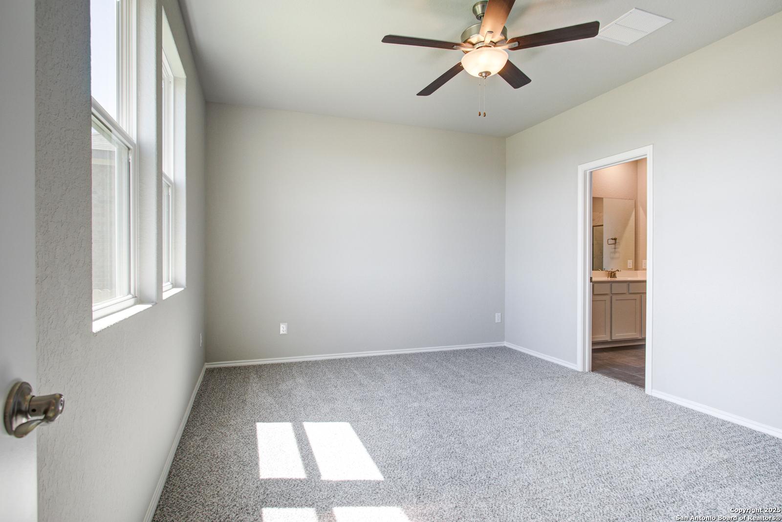 Bright bedroom with ceiling fan, gray carpet, large windows, and en-suite bathroom in Davidson Homes The Douglas G, San Antonio