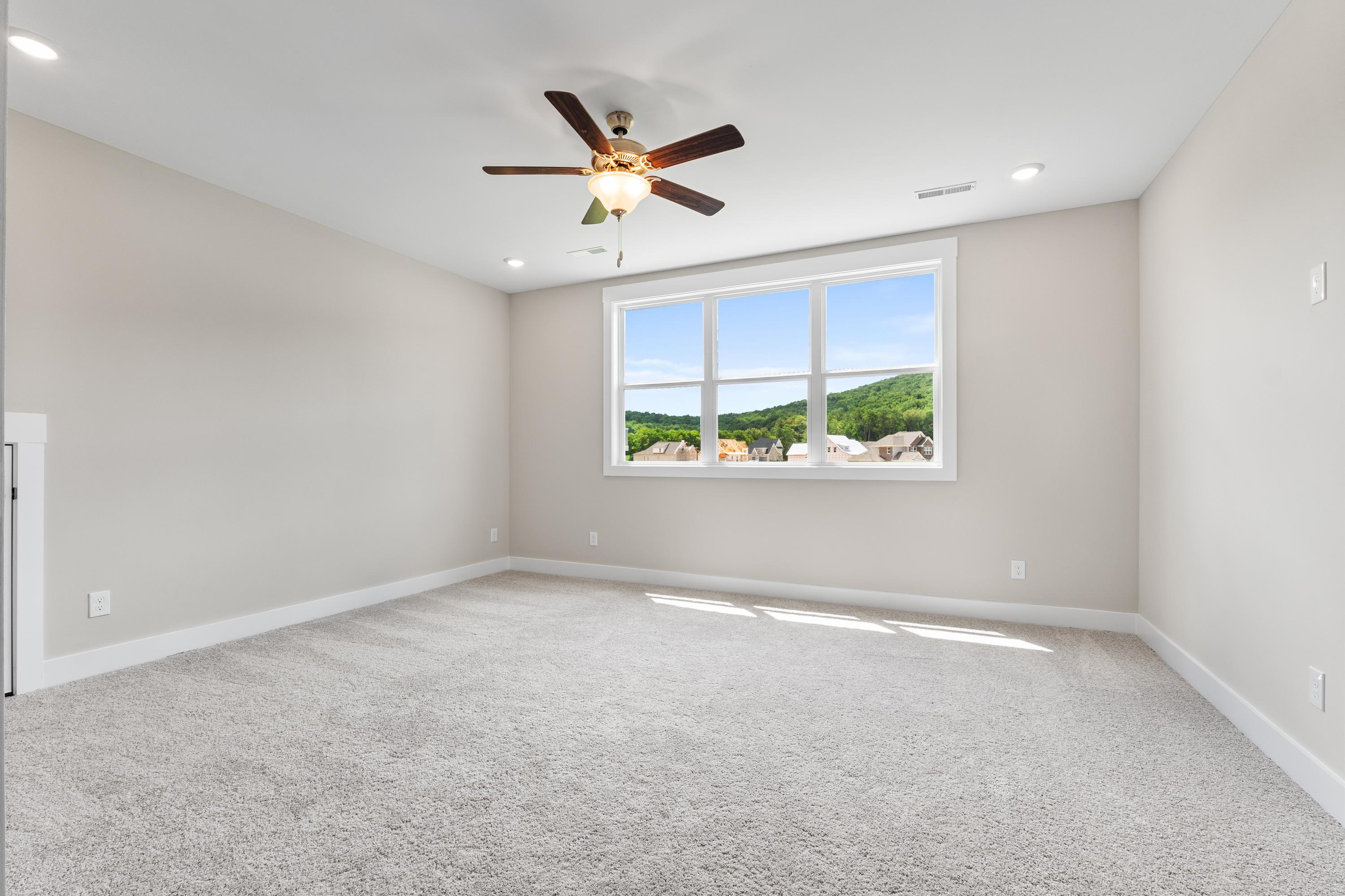 Spacious empty bedroom in The Oxford A with beige walls, carpeted floor, ceiling fan, and large window overlooking green hills