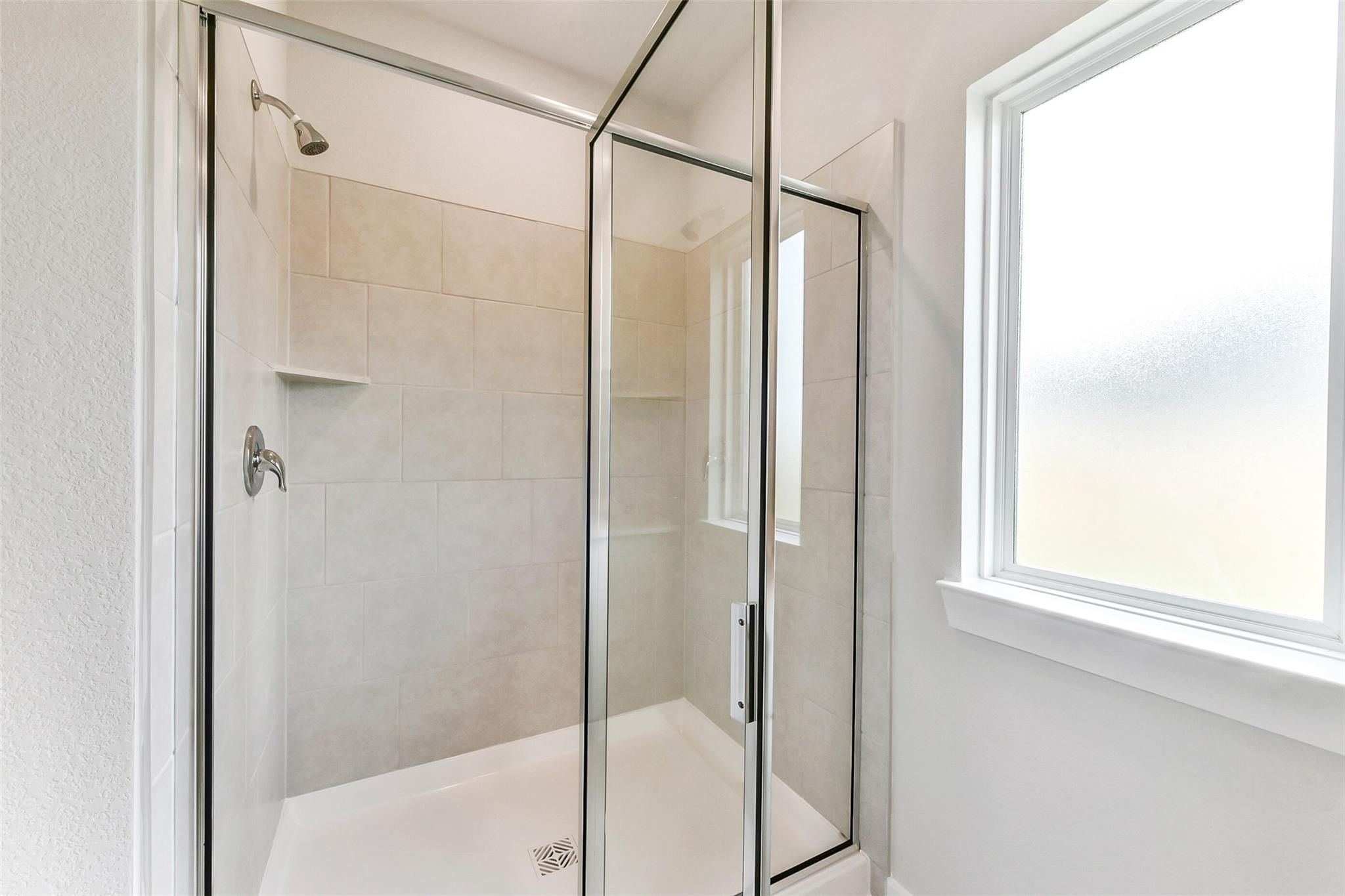 Frameless glass walk-in shower with beige tile surround, rainfall showerhead, and frosted window in The Costa B master bath, Dayton, Texas