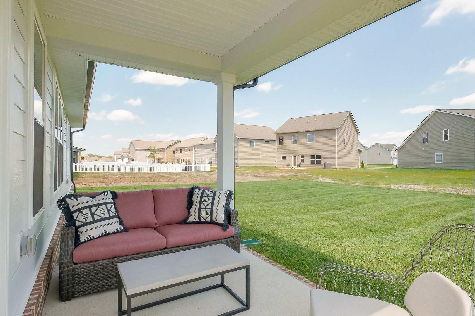 Covered patio at The Meadows in Smyrna TN featuring red sofa with patterned pillows, wicker chair, metal table, lush green lawn, and neighborhood homes