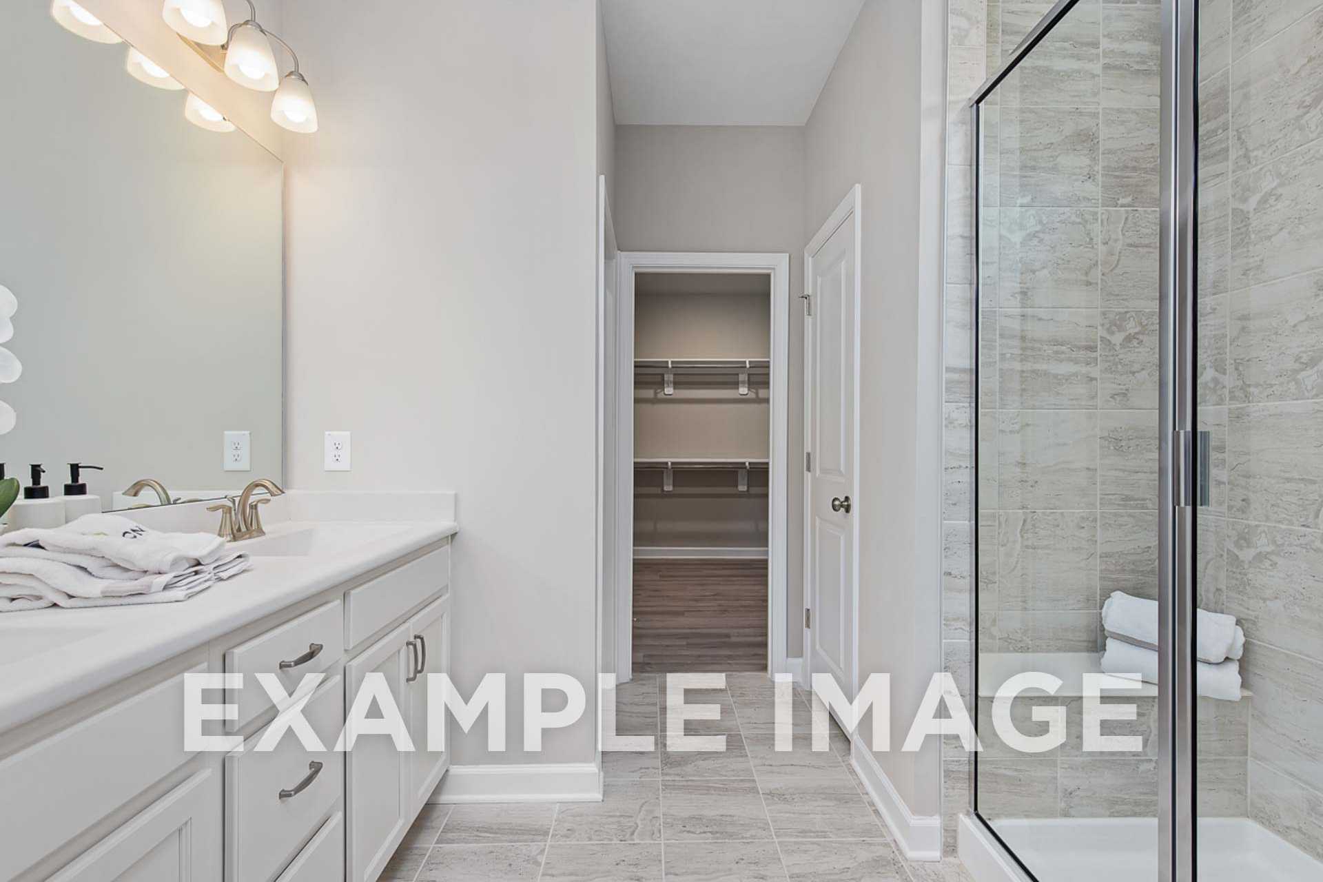 The Ash E master bathroom with white vanity, lighted mirror, frameless glass shower, and walk-in closet access