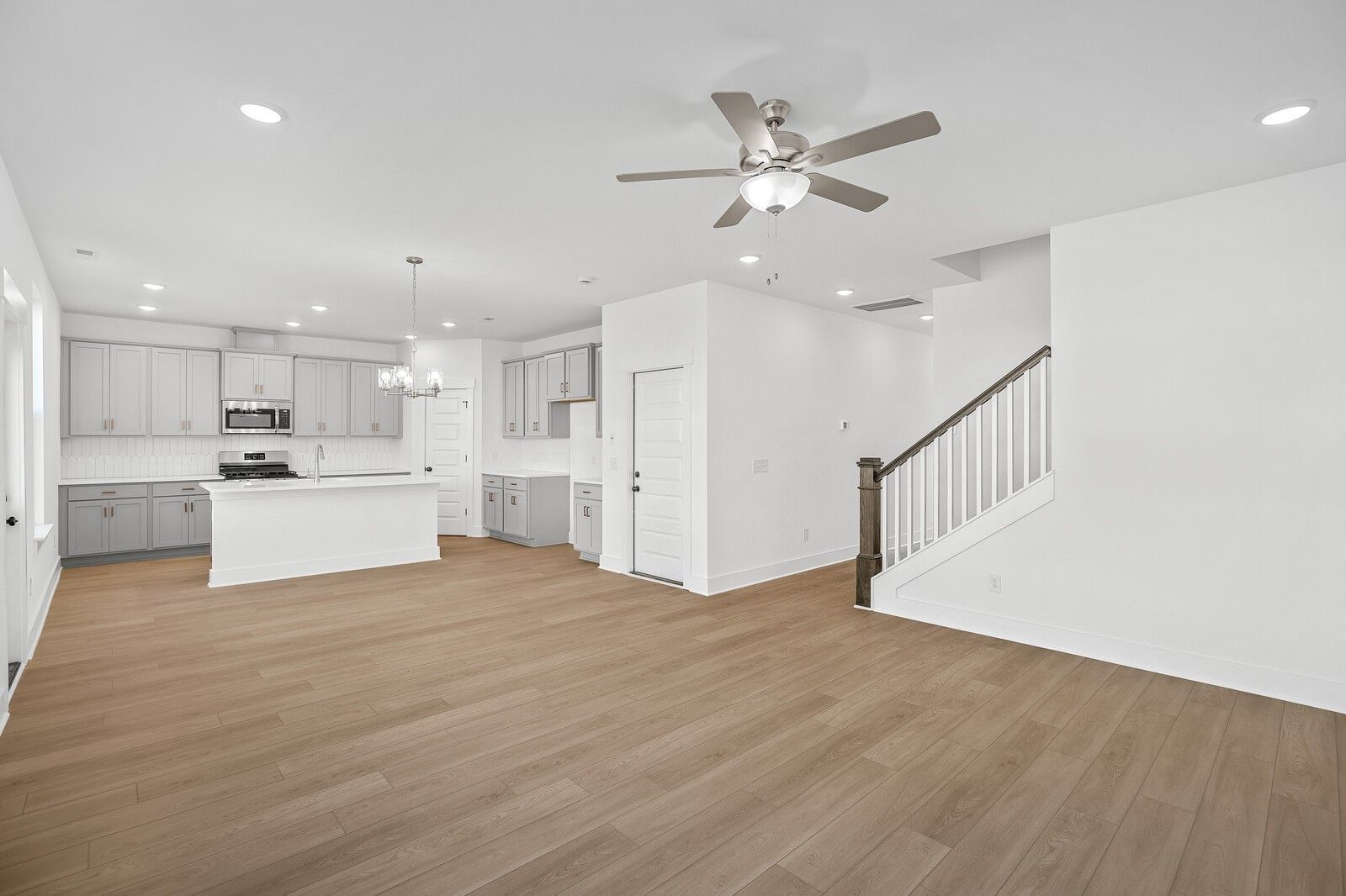 Open-concept kitchen with white cabinets, stainless appliances, and island flowing into living area with ceiling fan and staircase in Davidson Homes The Logan C, Gallatin TN