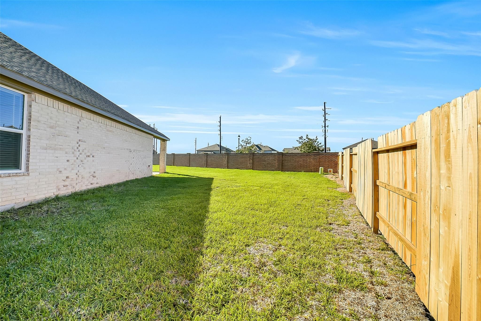 Side view of beige brick single-story home with green lawn, wooden privacy fence, and blue sky in Sunterra, Katy, Texas