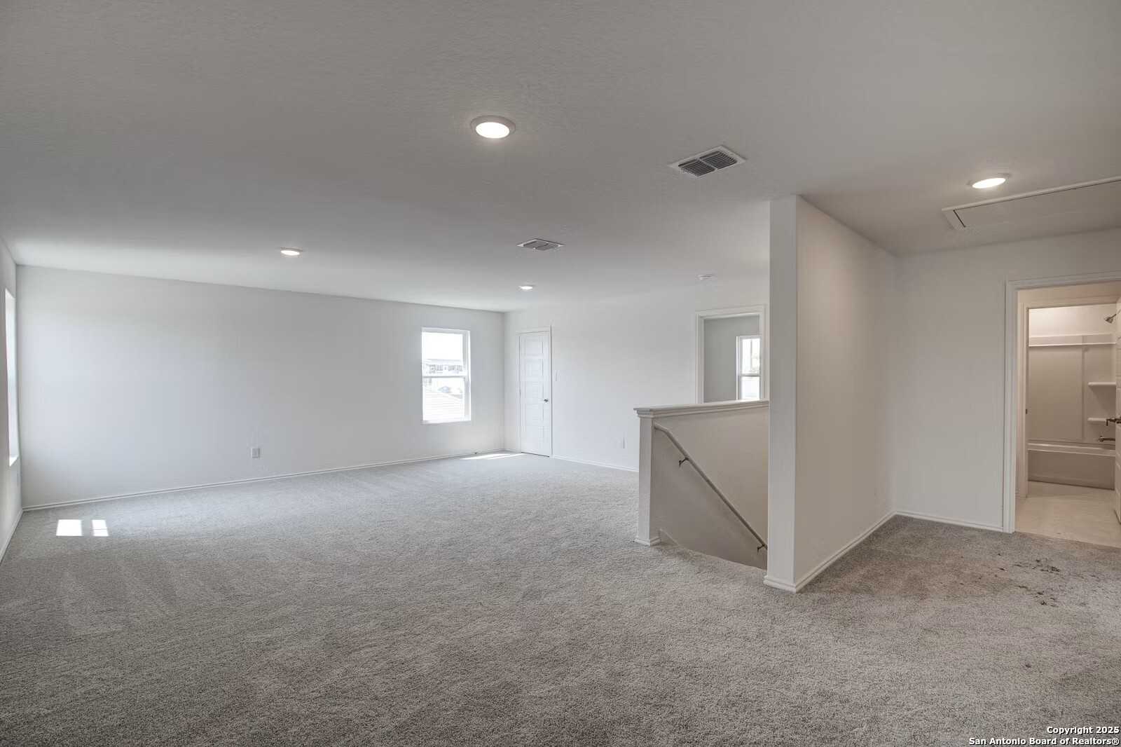 Spacious upstairs loft with gray carpet, white walls, staircase railing, and adjacent bathroom in The Douglas B, Hannah Heights, Seguin, Texas