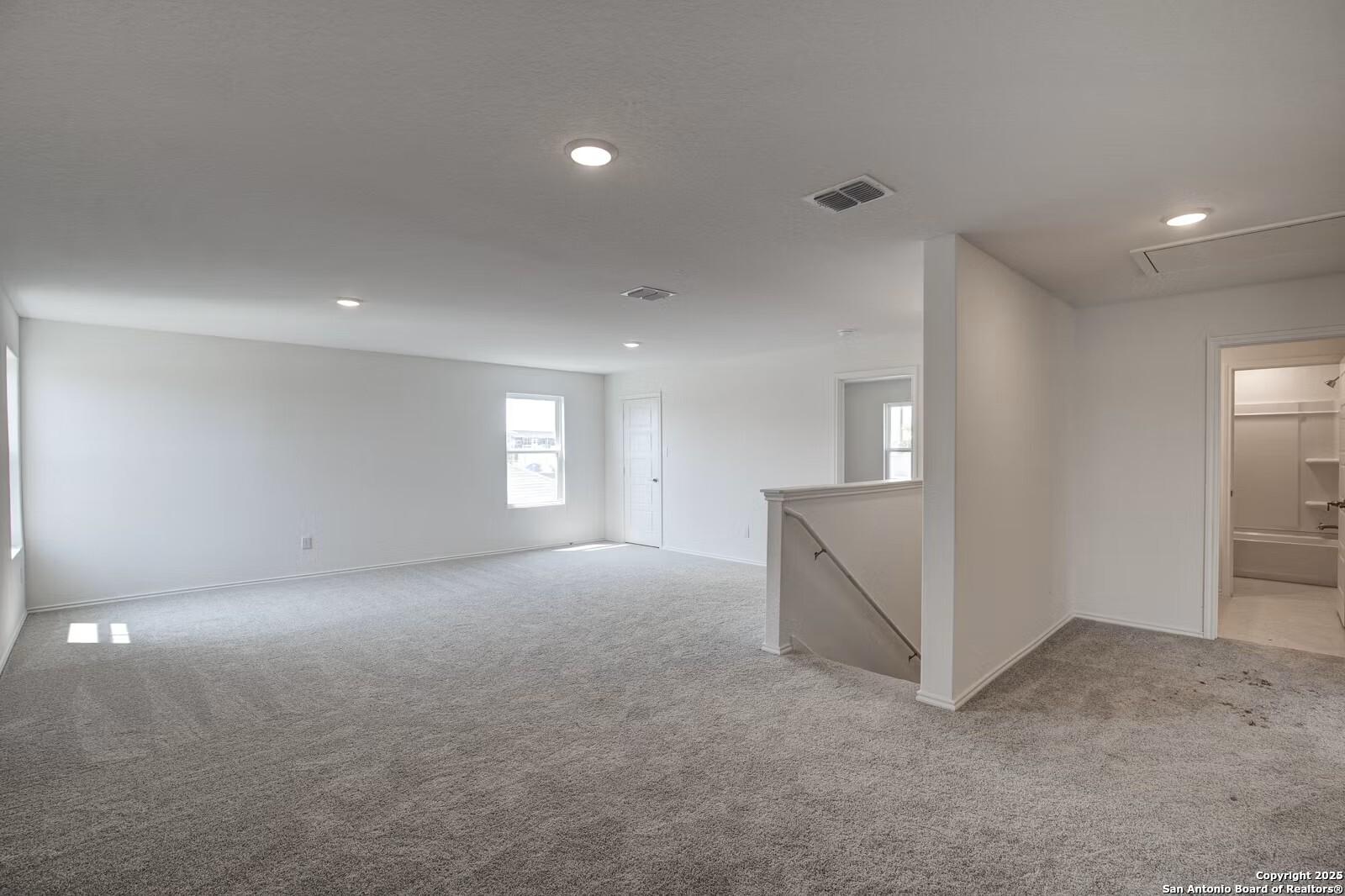 Spacious upstairs loft with gray carpet, white walls, staircase railing, and adjacent bathroom in The Douglas B, Hannah Heights, Seguin, Texas
