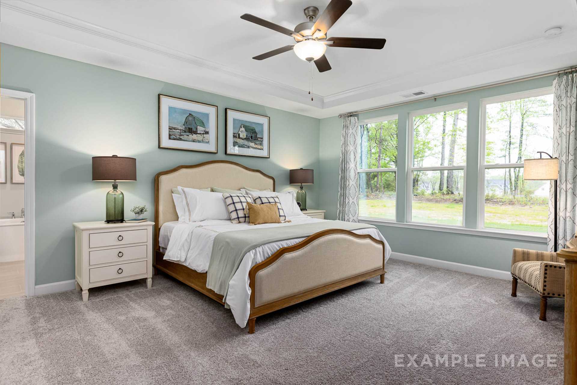 Spacious primary bedroom at Prince Place in Fuquay-Varina NC with tufted beige bed, ceiling fan, large windows, and artwork