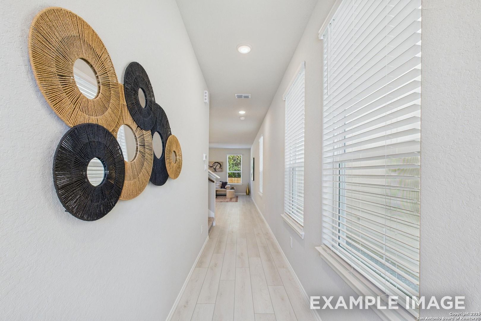 Bright hallway with woven circular wall art, light oak floors, and window blinds in Davidson Homes The Charlotte B, San Antonio, Texas