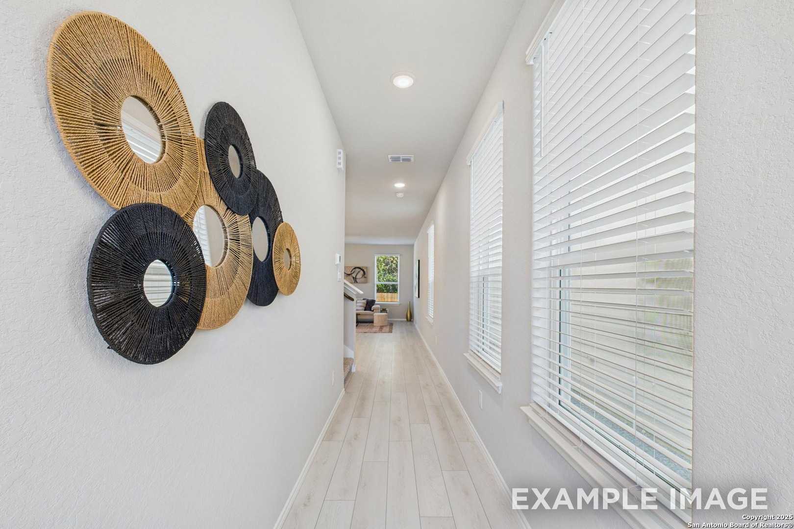 Bright hallway with woven circular wall art, light oak floors, and window blinds in Davidson Homes The Charlotte B, San Antonio, Texas