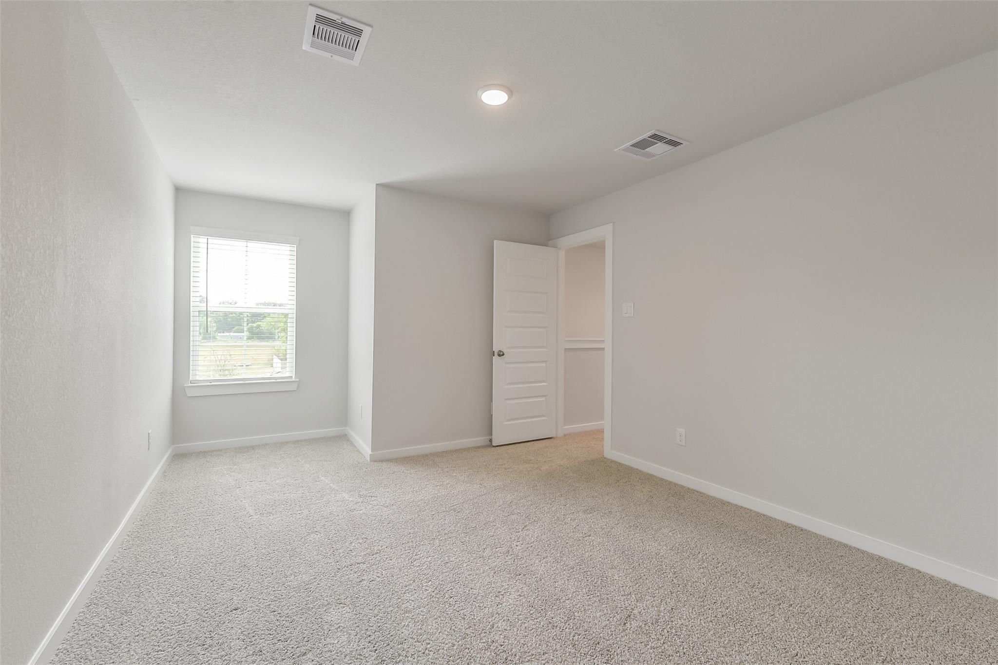 Bright secondary bedroom featuring light gray walls, beige carpet, window with blinds overlooking greenery in The San Marcos E, Conroe Texas