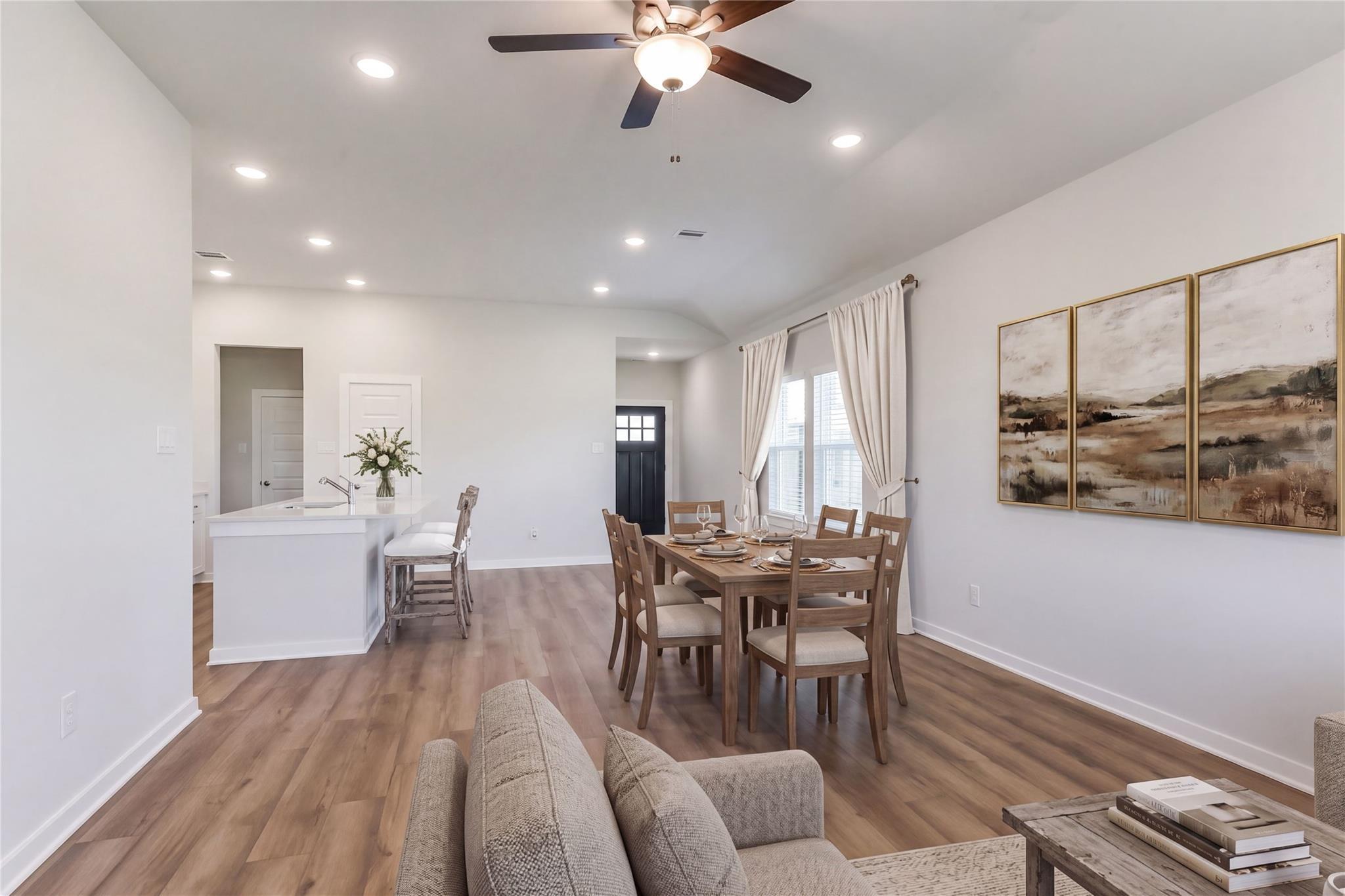 Open-concept living and dining area with white island, wooden table, beige sofa, ceiling fan, and abstract art in Davidson Homes The Costa C, Dayton, Texas
