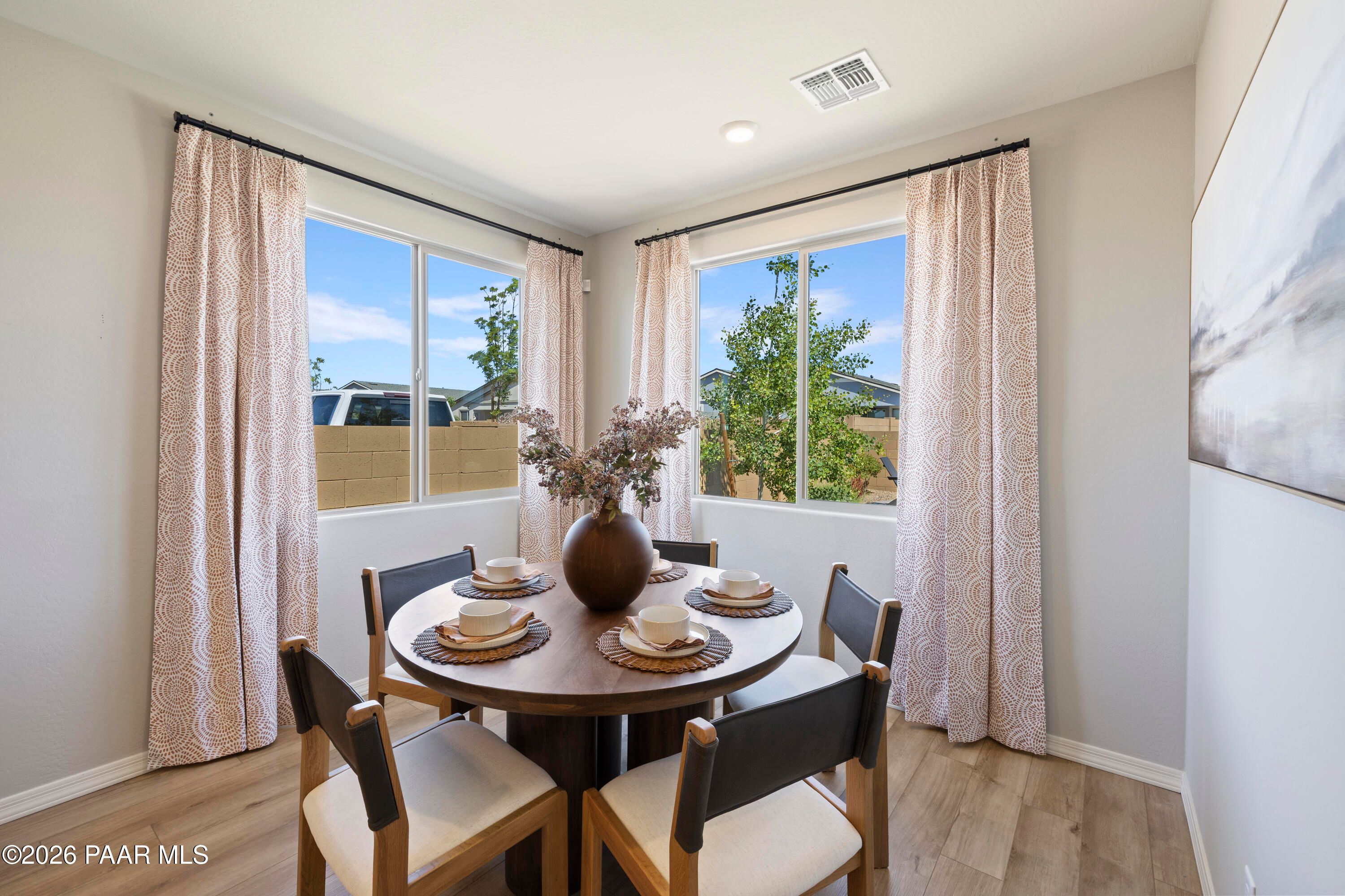 Elegant round dining table with set places and floral vase, large windows with beige curtains overlooking greenery in The Harmony A, Prescott Valley home