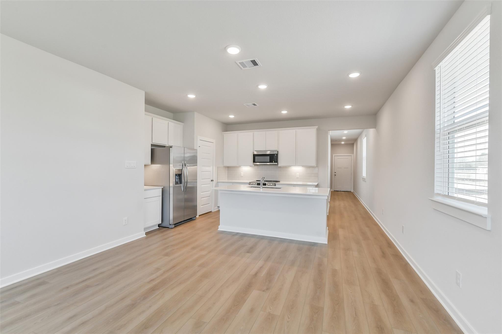 Modern open kitchen with white shaker cabinets, stainless fridge, large center island, and wood floors in Davidson Homes The Colorado F, Cleveland, Texas