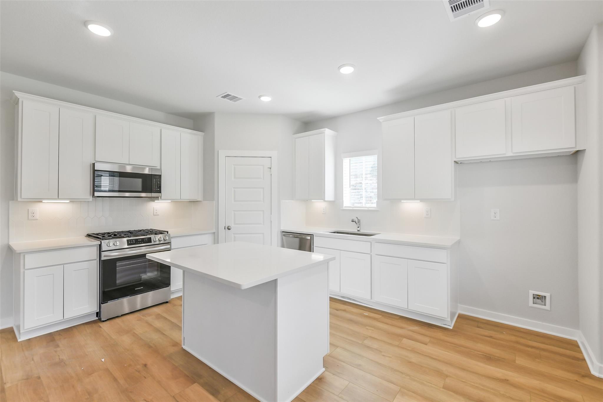 Bright white kitchen with stainless steel appliances, large center island, and hardwood floors in Davidson Homes The Trinity F, Magnolia, Texas