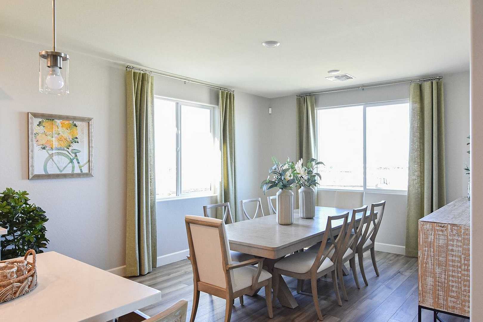Spacious dining room in The Summit B home with wooden table, upholstered chairs, floral vase, plants, and open kitchen view