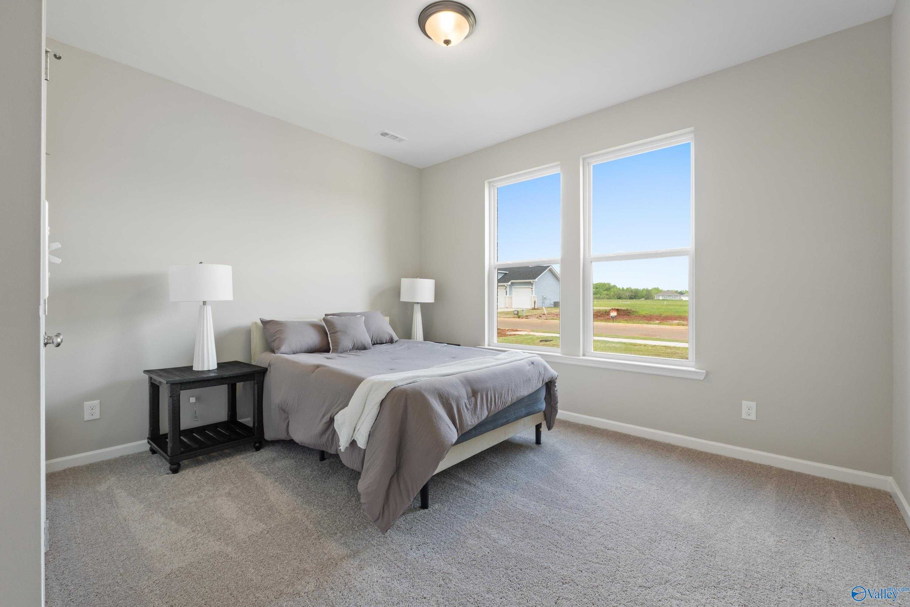Cozy secondary bedroom with queen bed, gray bedding, lamps, and large windows showing rural view in Davidson Homes The Luna, Hazel Green, AL