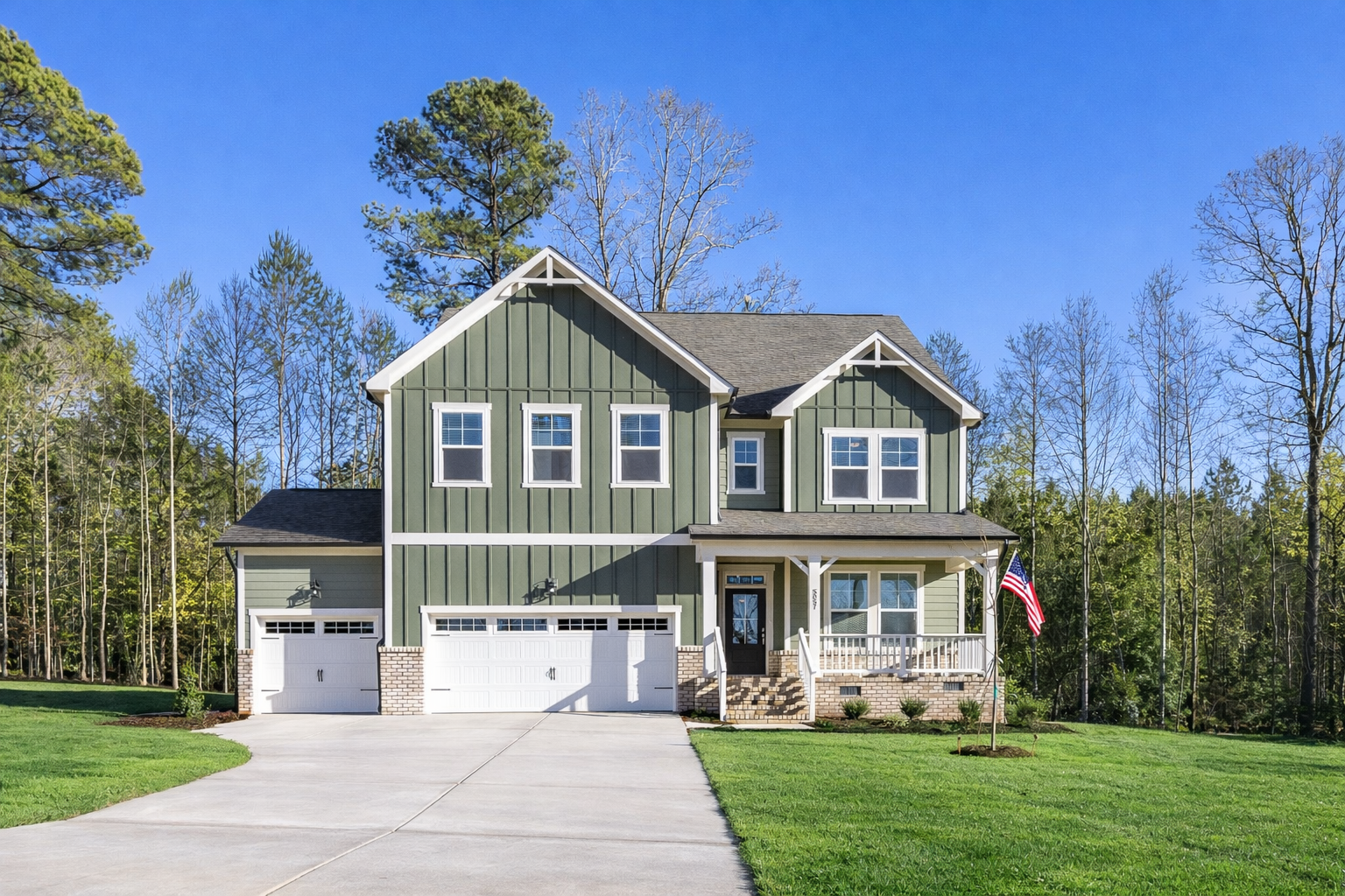 Two-story sage green craftsman home at Rockport in Wake Forest NC with covered front porch, American flag, and two-car garage amid pine trees