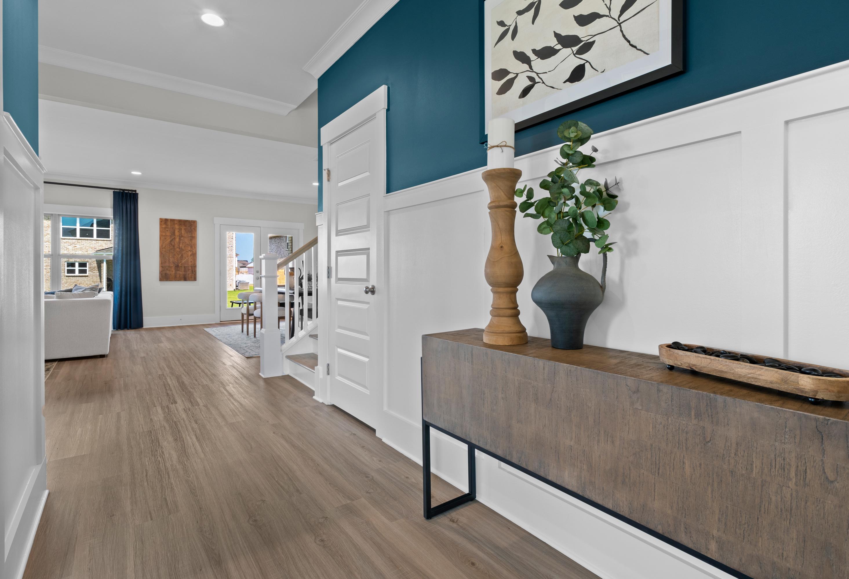 Spacious foyer in The Chelsea A with teal accent wall, white wainscoting, hardwood floors, console table, vase, and staircase