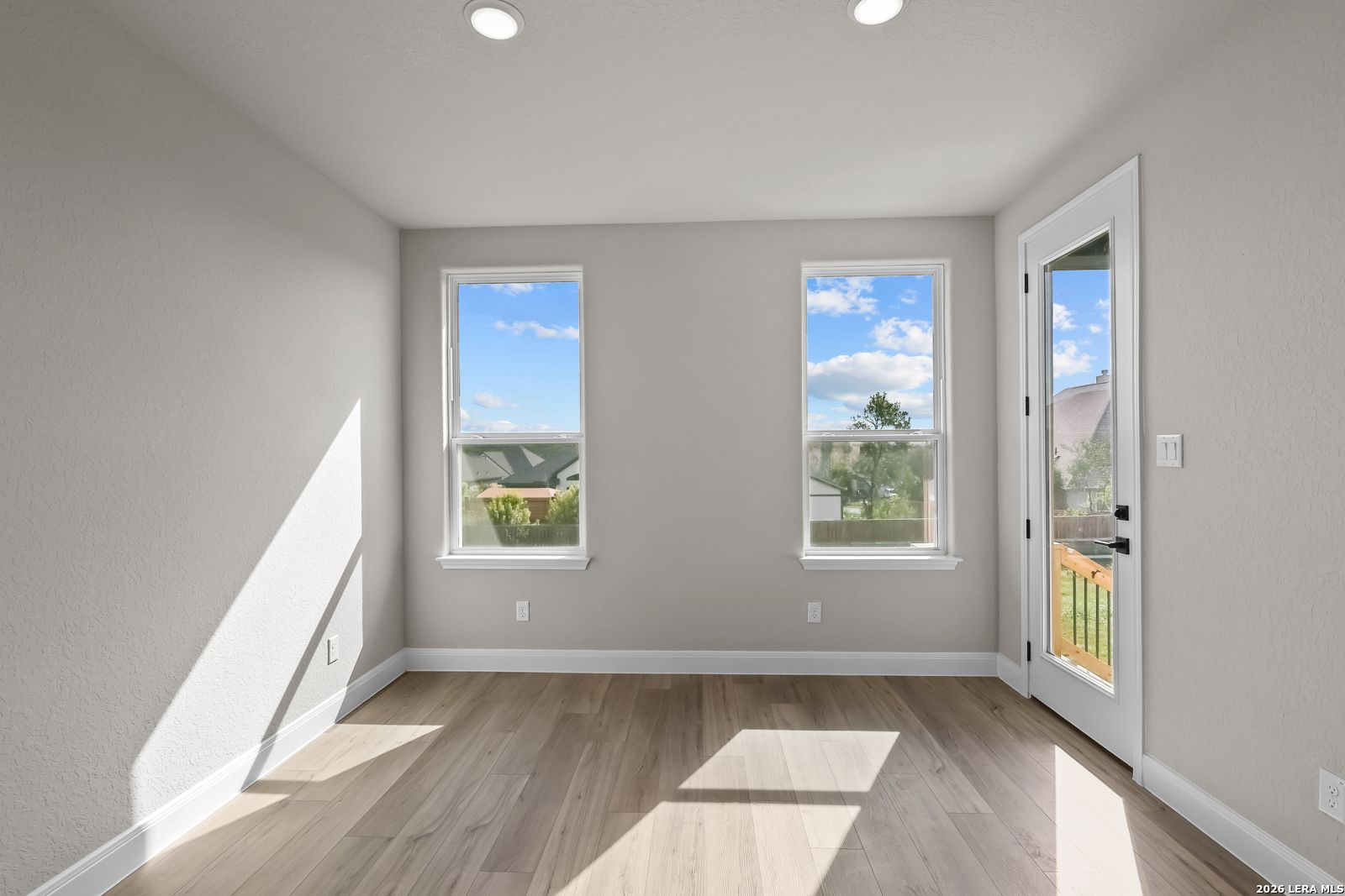Bright empty room with wood-look flooring, two windows, and glass door to backyard in Davidson Homes The Foster E, Castroville, TX