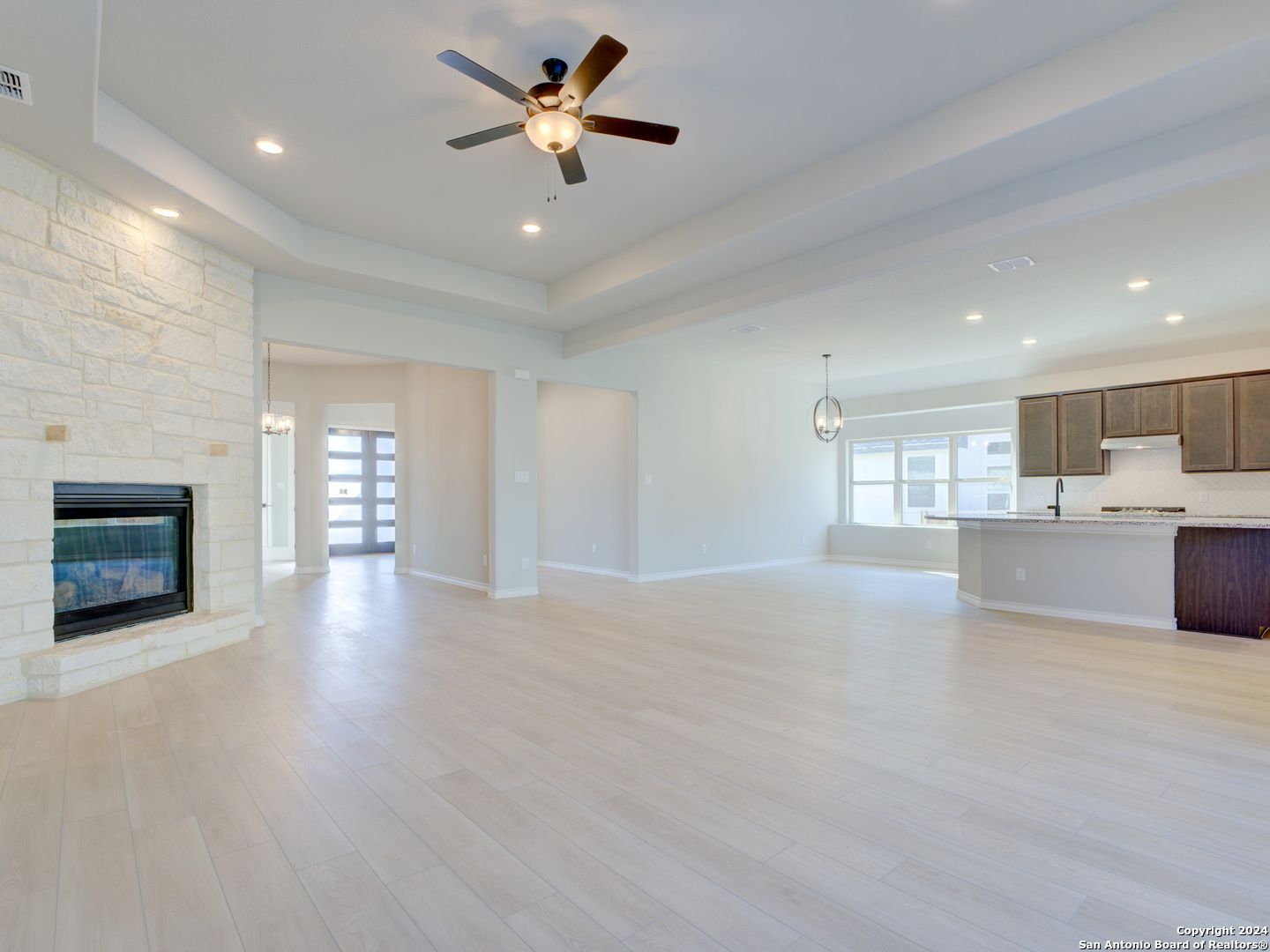 Spacious open-concept living room with stone gas fireplace, ceiling fan, and adjacent kitchen in 4-bedroom The Summerlin A home, Castroville, Texas