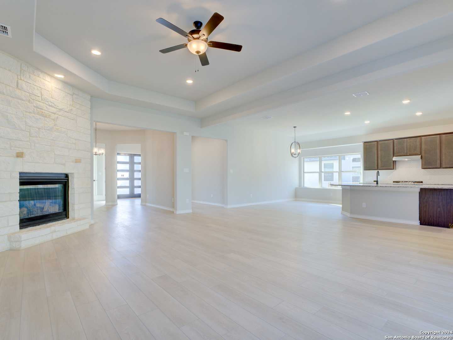 Spacious open-concept living room with stone gas fireplace, ceiling fan, and adjacent kitchen in 4-bedroom The Summerlin A home, Castroville, Texas