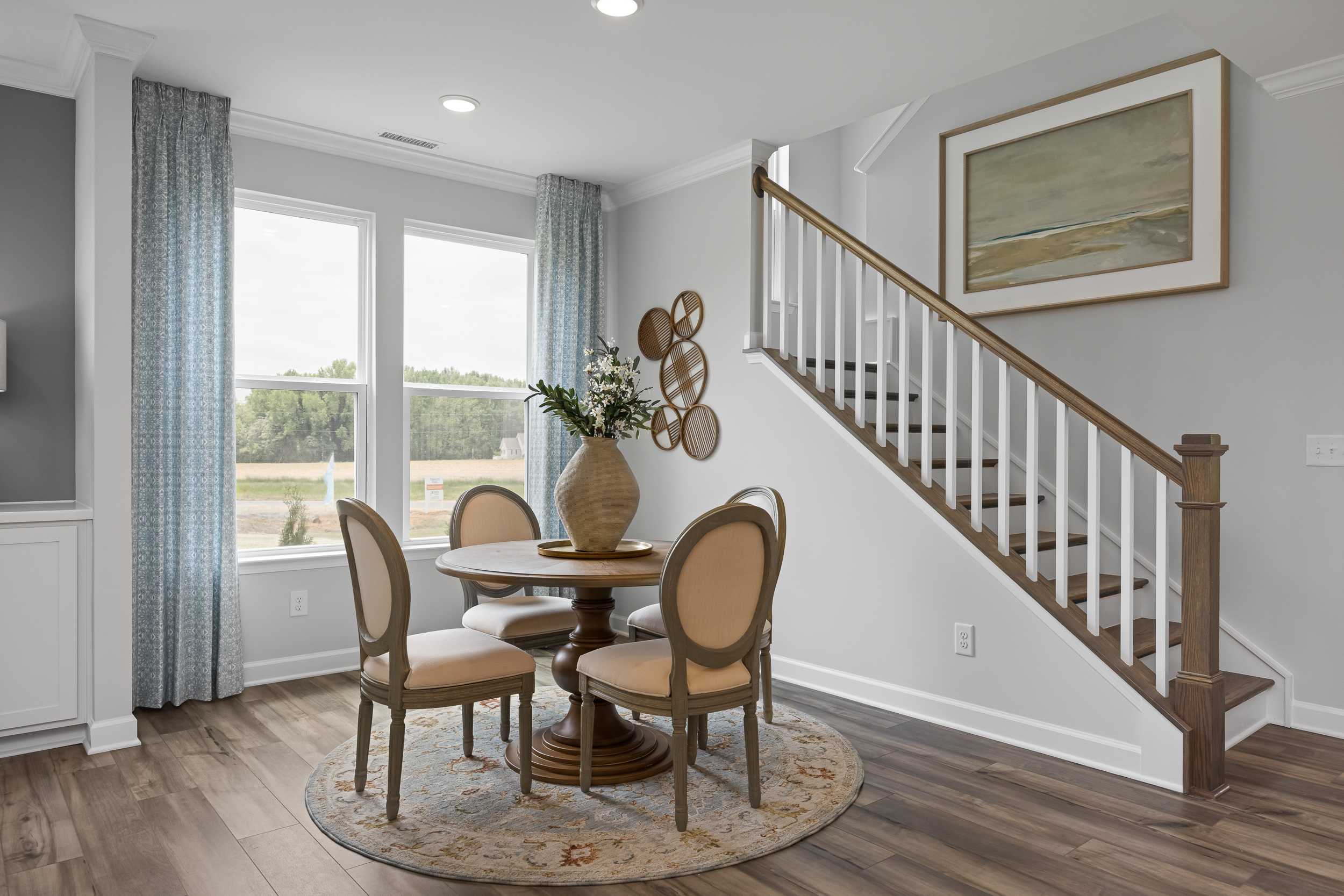 Cozy dining nook in The Magnolia A with round table, upholstered chairs, floral vase, large windows, and white staircase