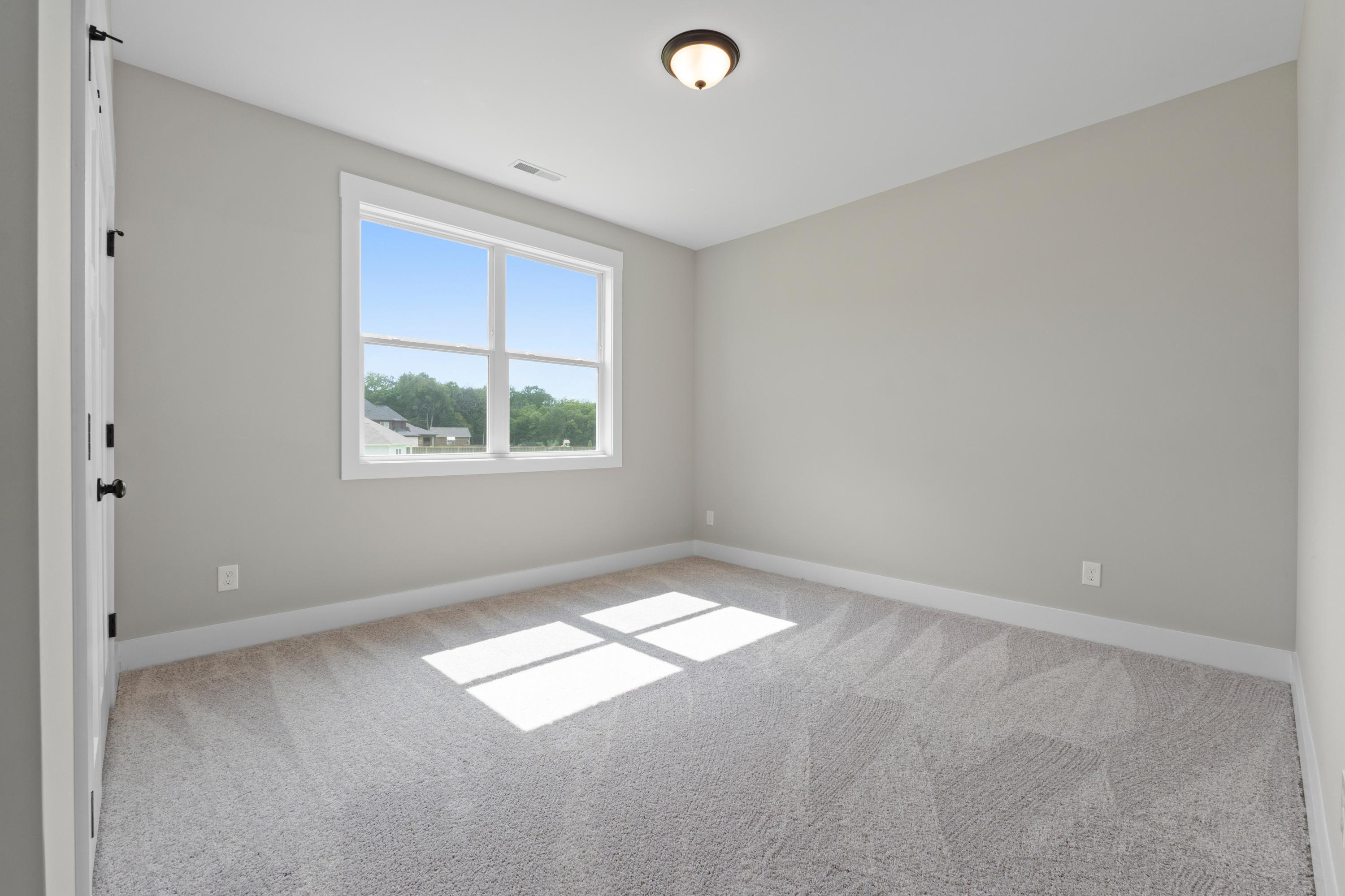 Bright secondary bedroom in The Oxford B with large sunny window, neutral gray walls, and plush carpet