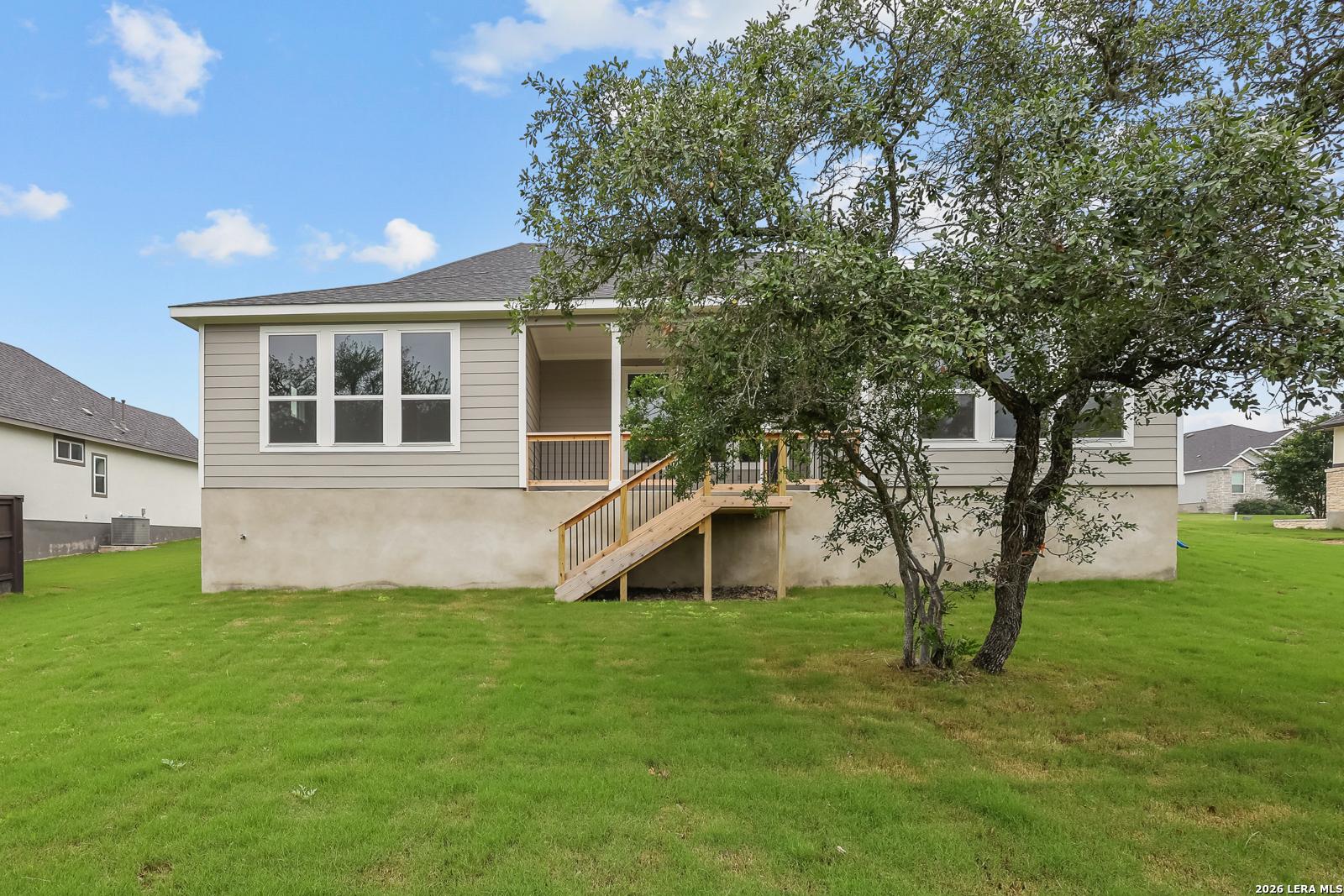 Back view of The Belmont H 4-bedroom home featuring covered porch with wooden stairs, mature oak tree, and lush green lawn in Potranco Oaks, Castroville, Texas