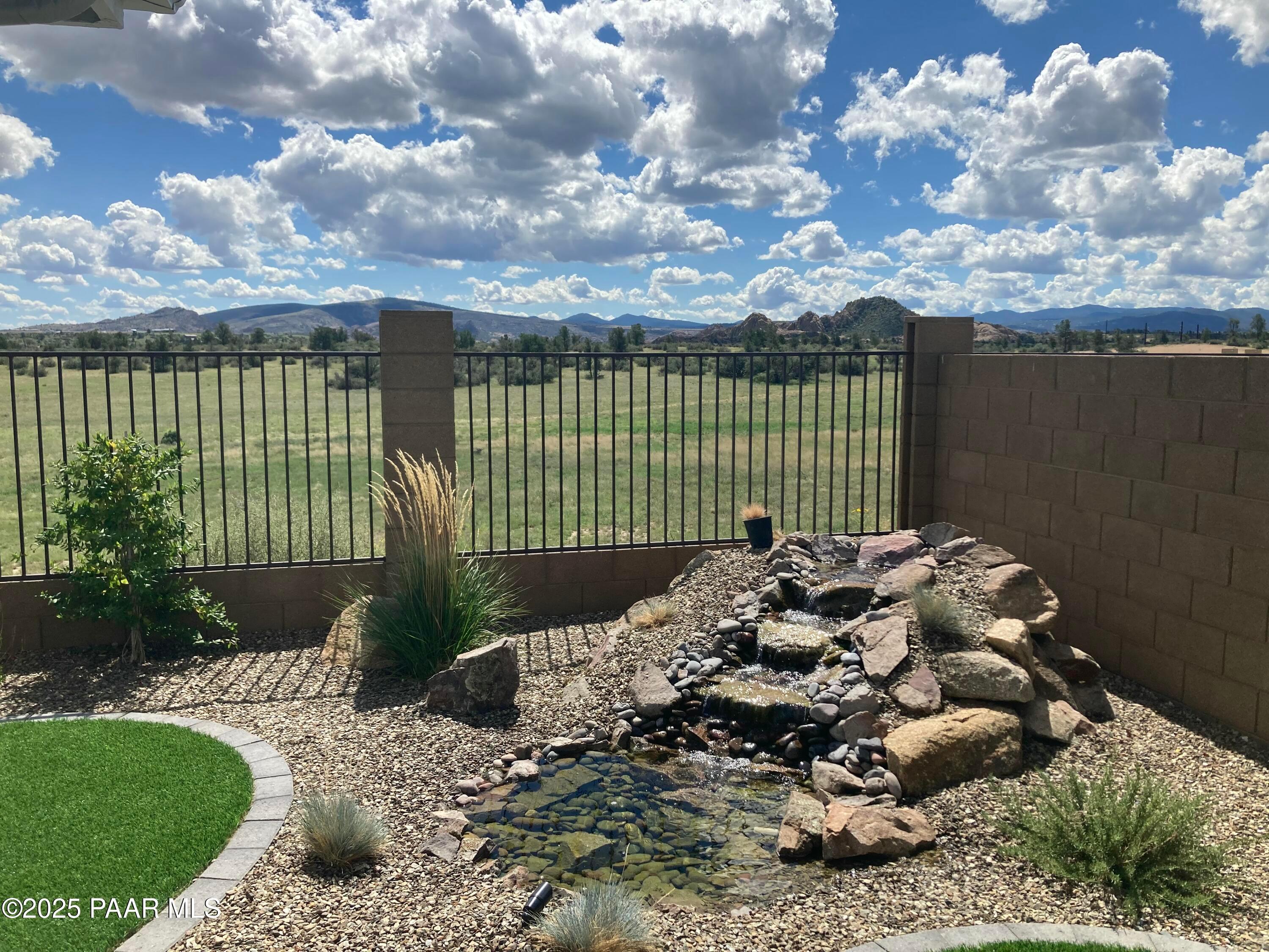 Tranquil rock waterfall cascading into pond amid gravel landscaping, circular turf, desert plants, fenced yard with mountain views in Prescott, Arizona