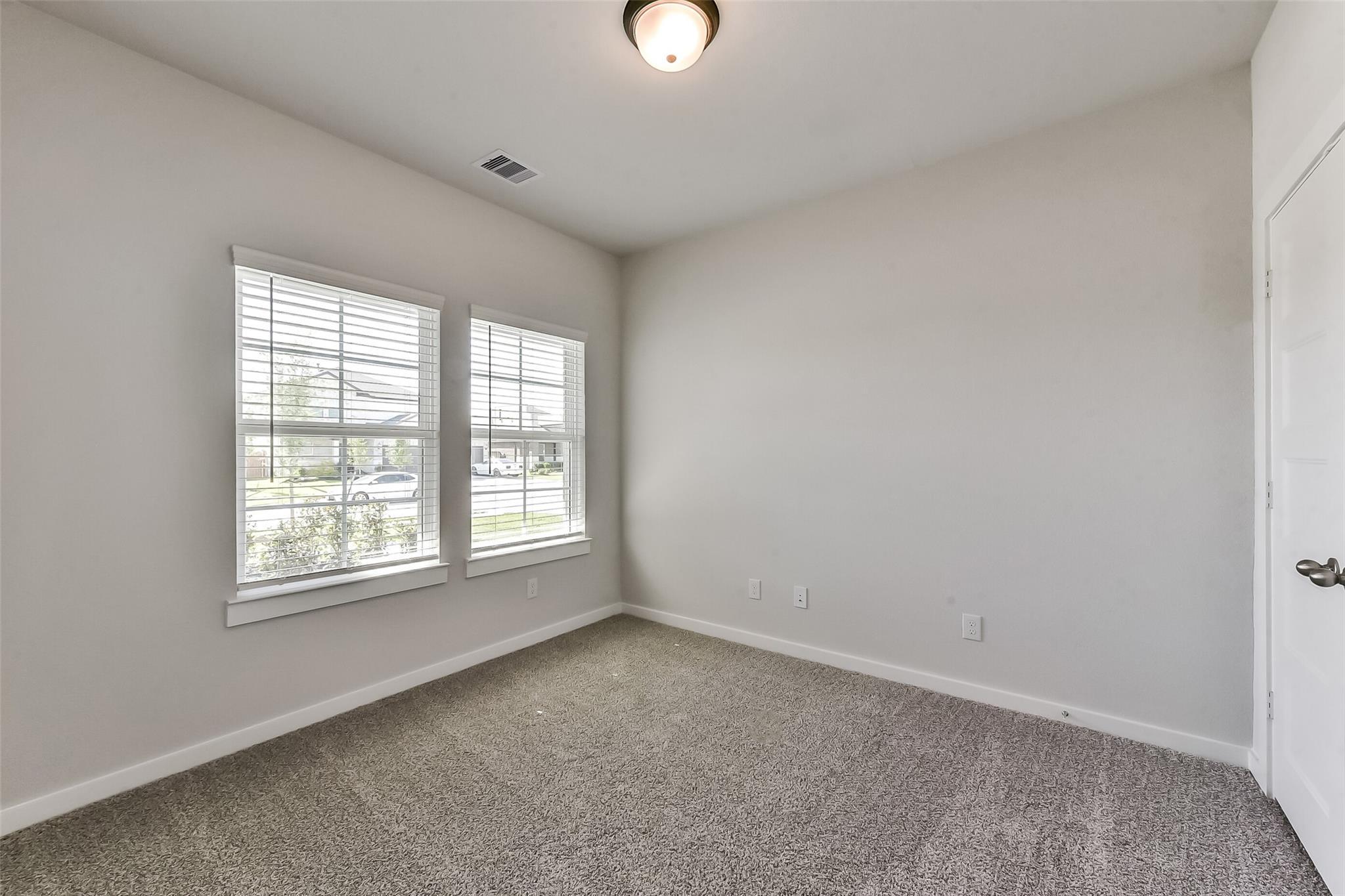Bright bedroom with beige walls, gray carpet, blinds on dual windows, and ceiling light in Davidson Homes The Costa C, Dayton, Texas