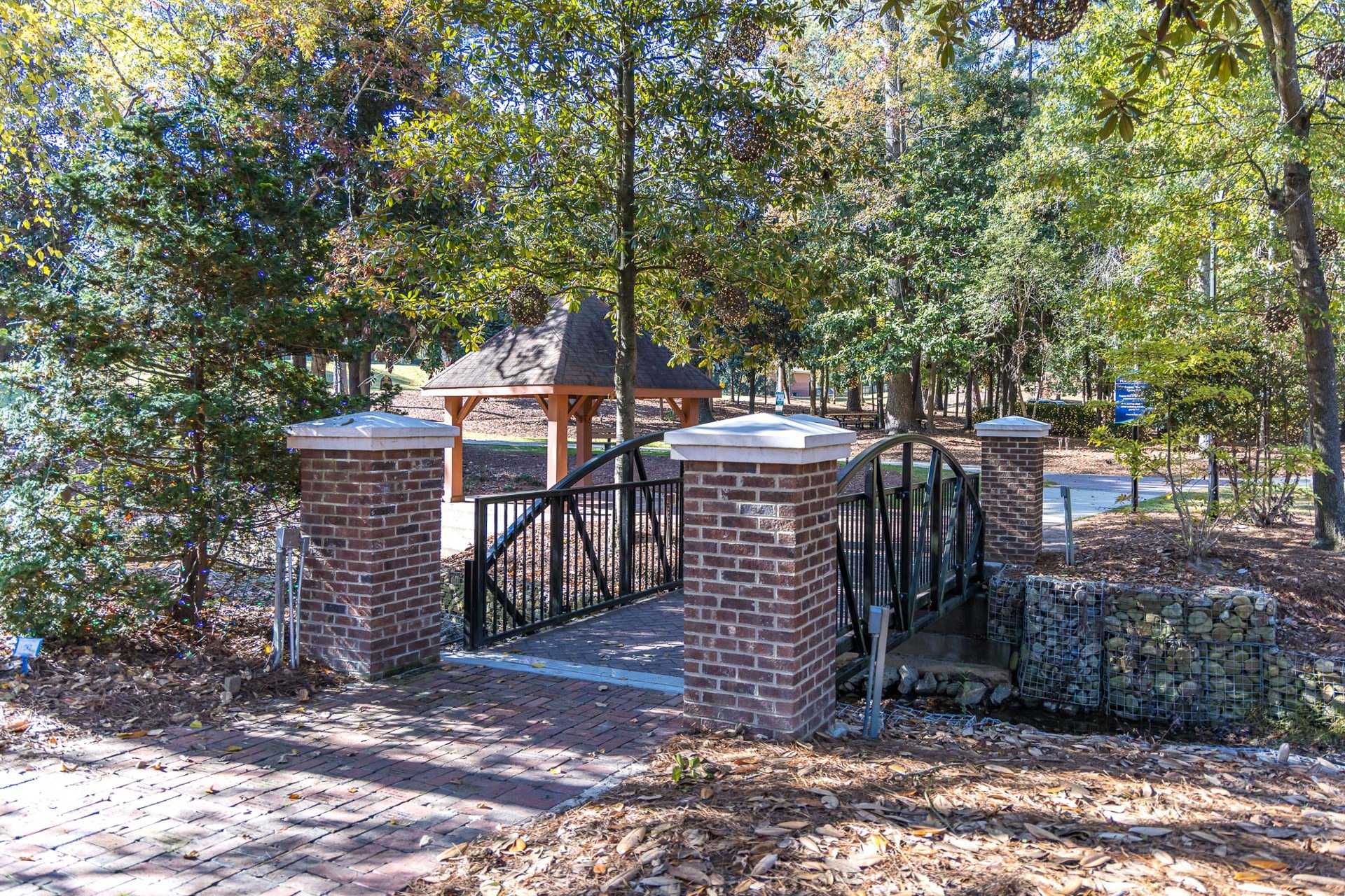 Pedestrian footbridge with brick pillars and wrought iron railing at Prince Place in Fuquay-Varina, NC amid wooded landscape