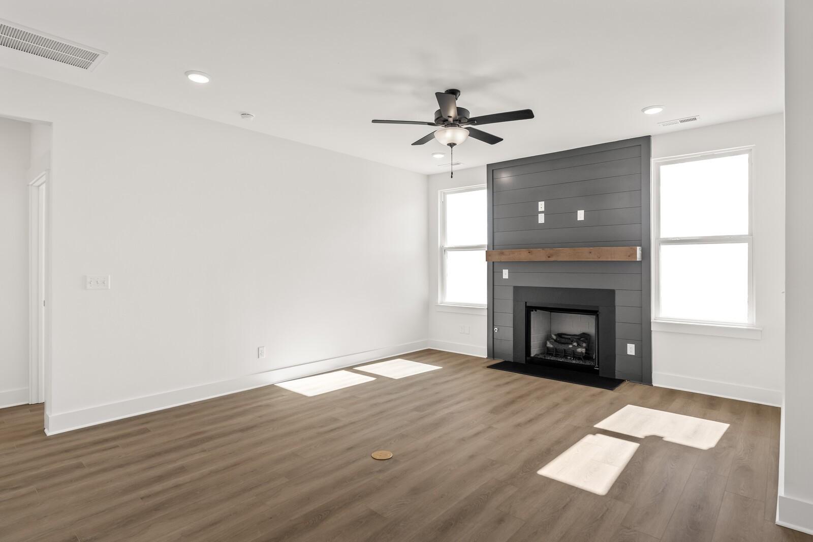Modern living room with gas fireplace, wood mantel, ceiling fan, large windows, and hardwood floors in Davidson Homes The Ash A, Gallatin, TN