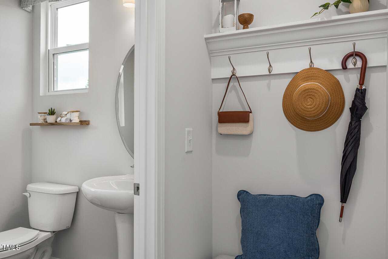 Cozy powder room featuring white toilet, pedestal sink, round mirror, plant shelves, and hooks with straw hat, umbrella, bag in Davidson Homes The Graham, Fuquay-Varina, NC