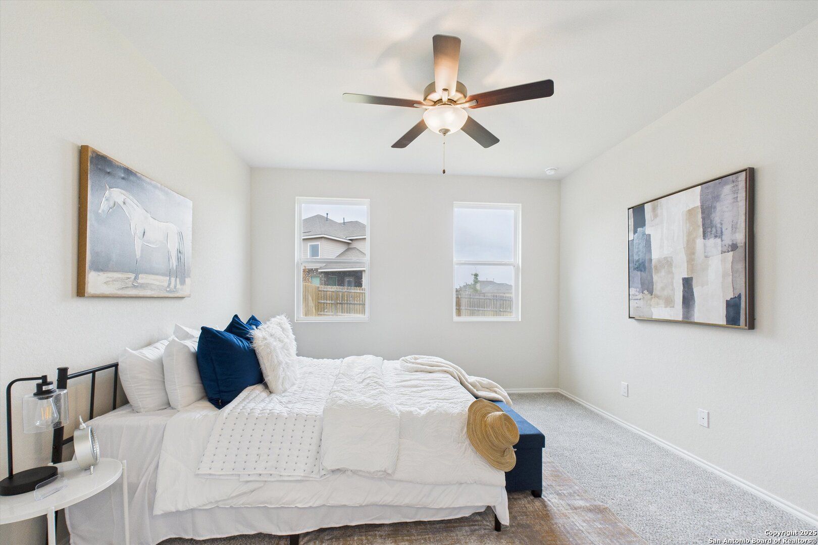Serene master bedroom with white duvet, blue pillows, ceiling fan, abstract art, and windows in Davidson Homes The Asheville K, Bricewood, San Antonio