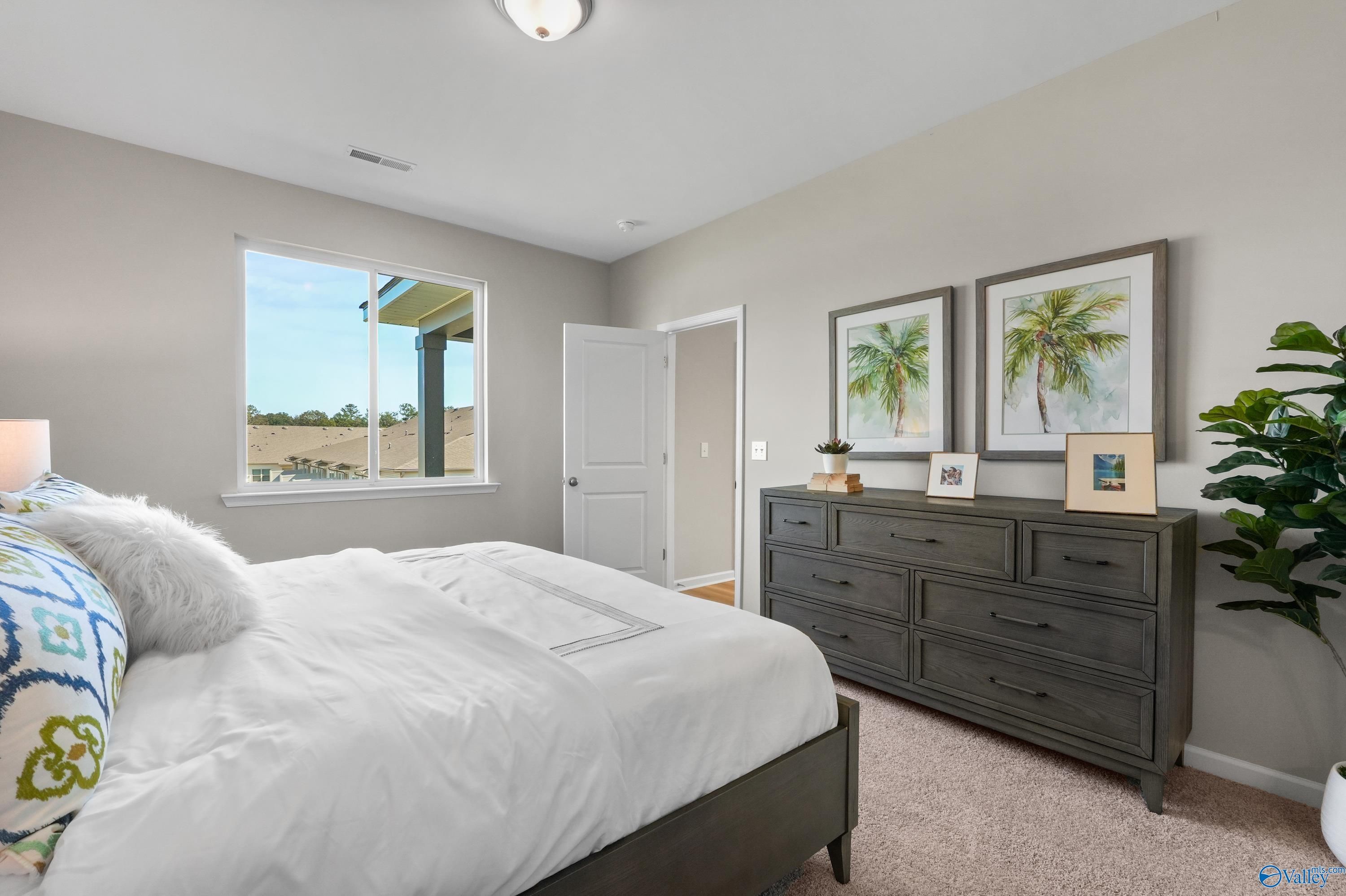 Serene master bedroom with king bed, plush bedding, dark dresser, palm art, and window view in Evermore Homes The Malibu, Madison AL