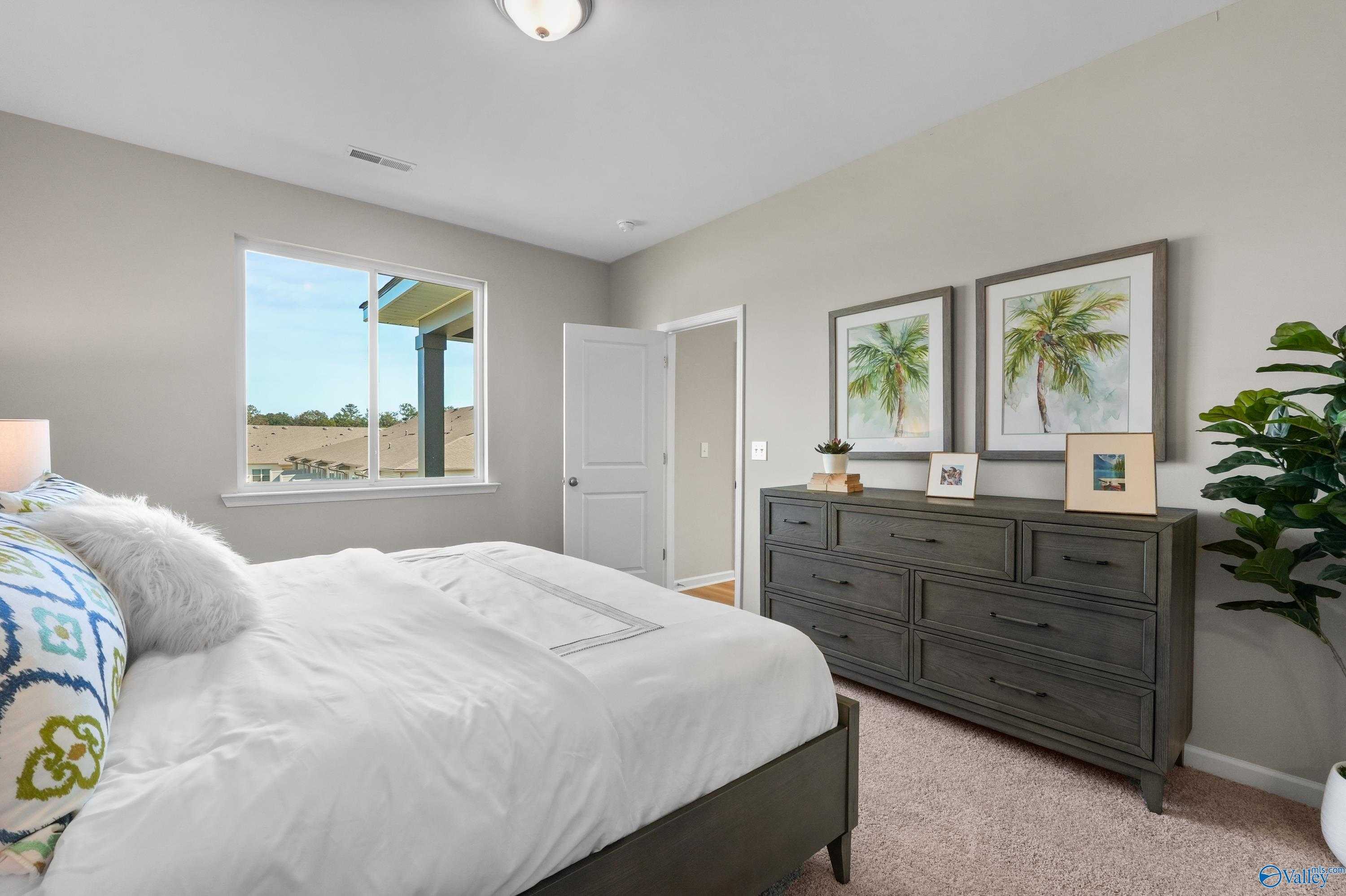 Serene master bedroom with king bed, plush bedding, dark dresser, palm art, and window view in Evermore Homes The Malibu, Madison AL