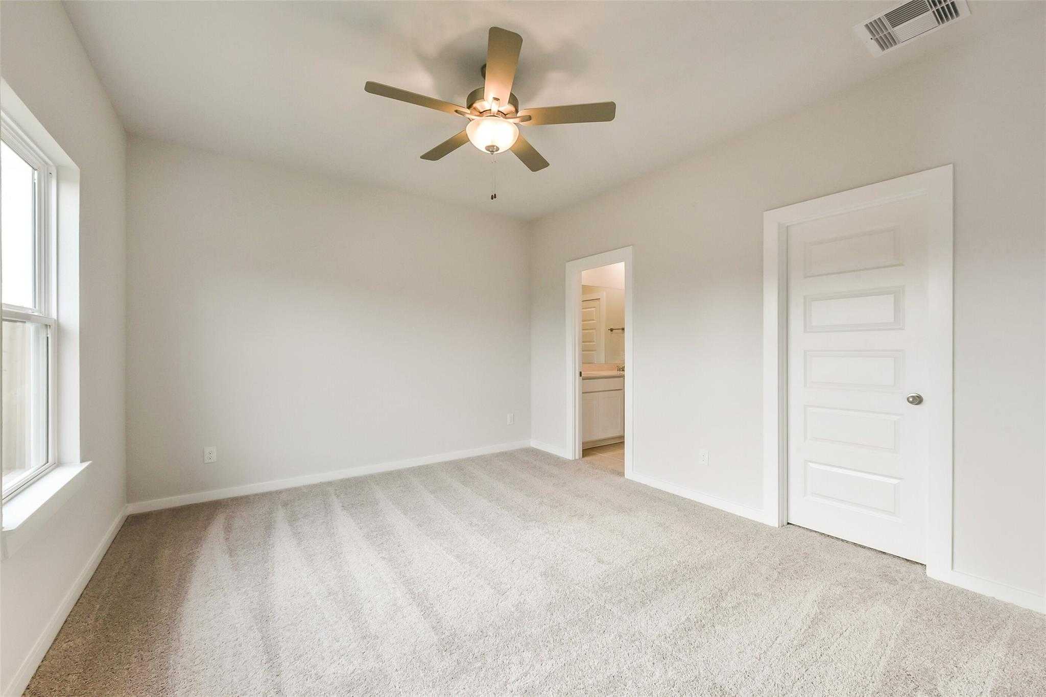Bright secondary bedroom with ceiling fan, beige carpet, large windows, and adjacent bathroom in Davidson Homes The Frio G, Dayton, Texas