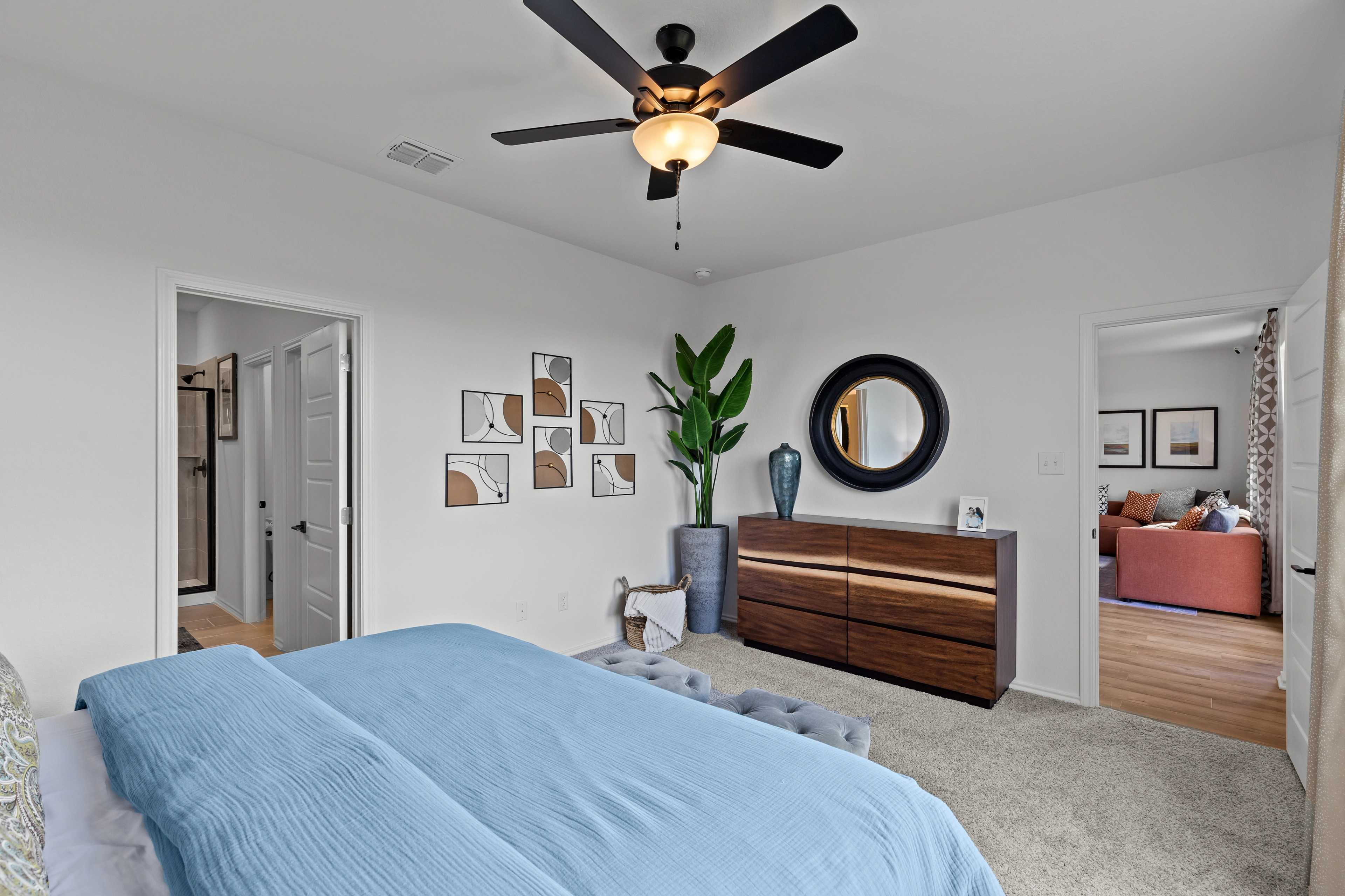 Spacious master bedroom in The Douglas C with blue upholstered bed, wooden dresser, round mirror, and potted plants