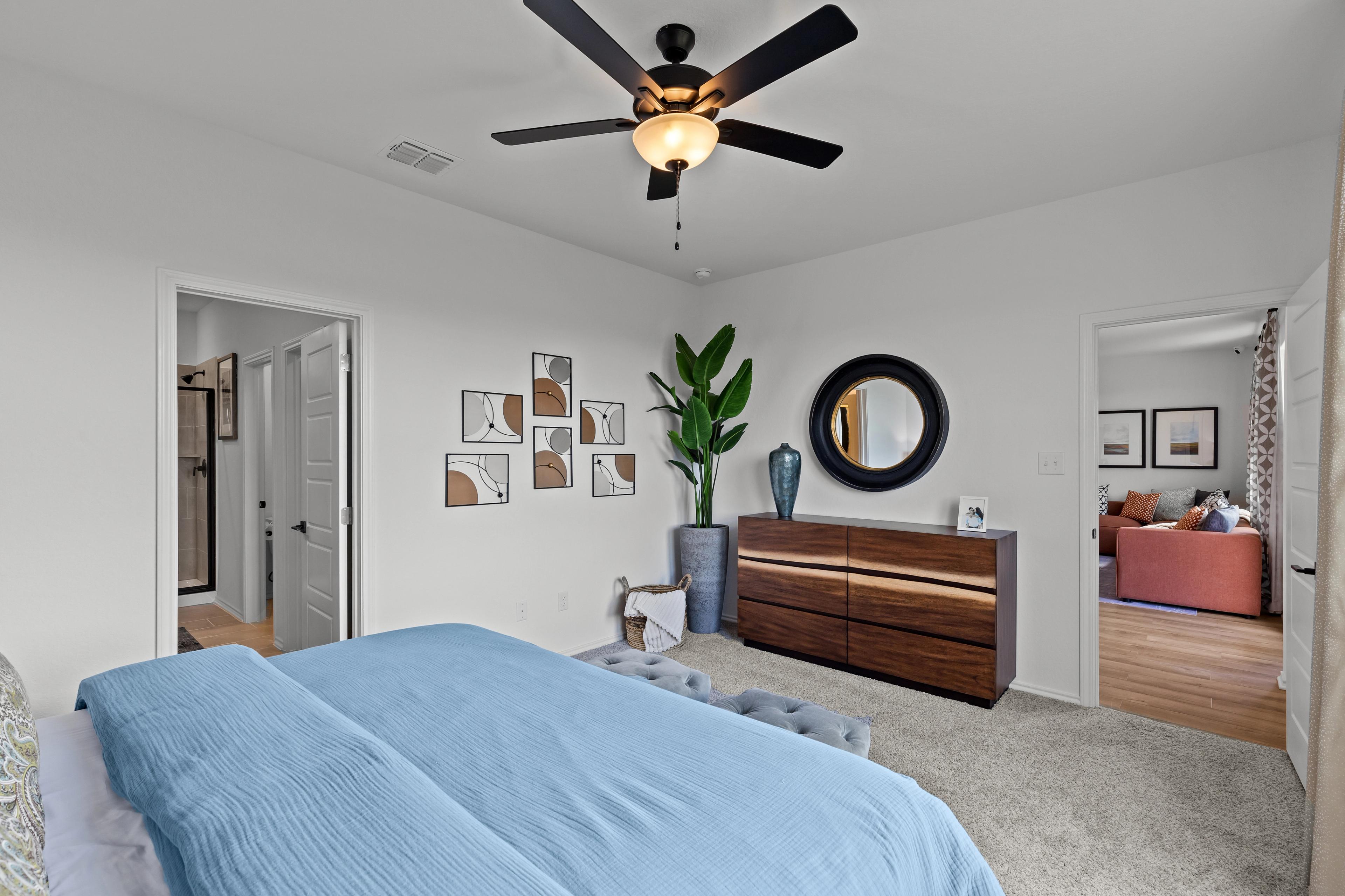 Spacious master bedroom in The Douglas F with blue tufted bed, walnut dresser, potted fern, round mirror, and ceiling fan