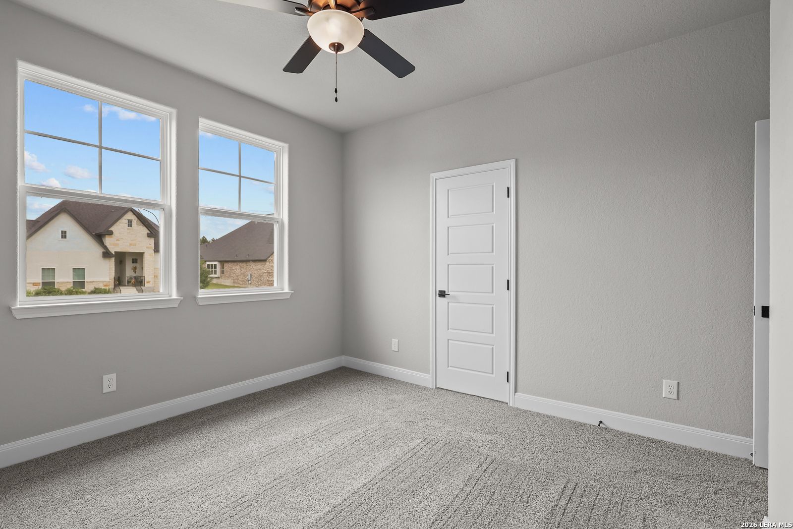 Bright empty bedroom with gray walls, large windows overlooking neighborhood, ceiling fan in The Belmont H, Castroville, Texas