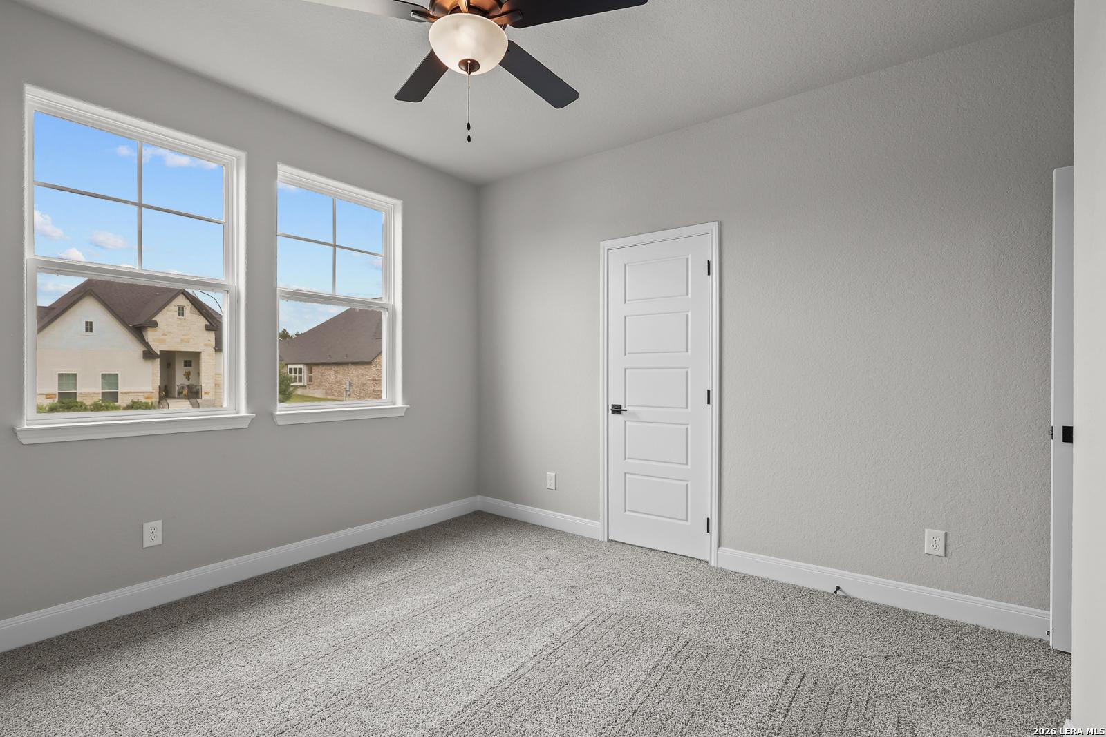 Bright empty bedroom with gray walls, large windows overlooking neighborhood, ceiling fan in The Belmont H, Castroville, Texas