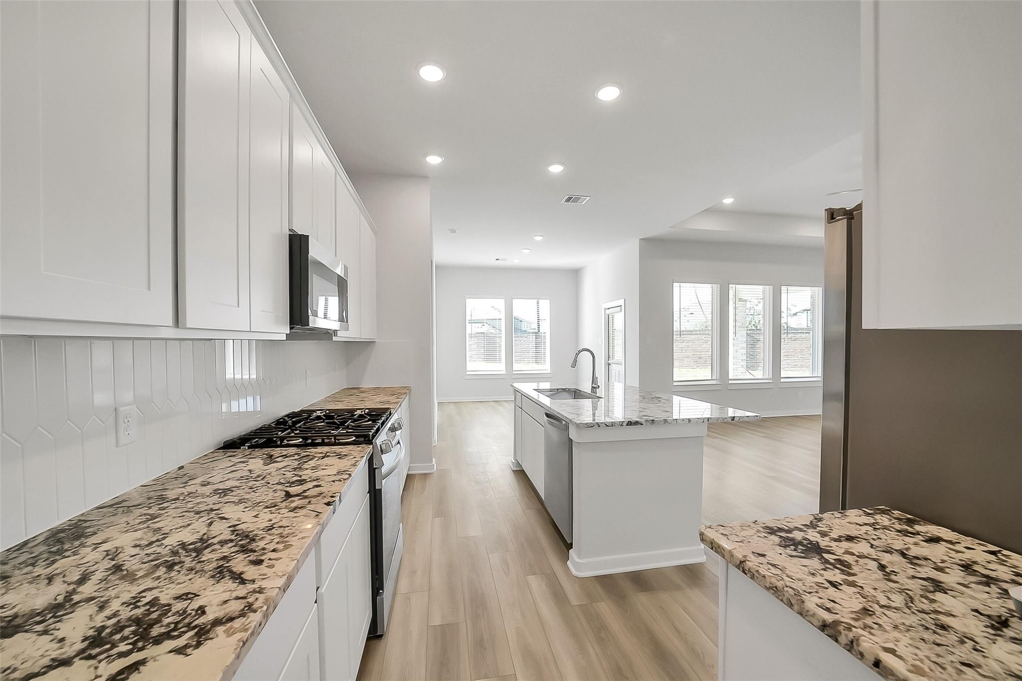 Open-concept kitchen with white shaker cabinets, speckled granite counters, stainless gas range, and island in Davidson Homes The Acadia A, Katy TX