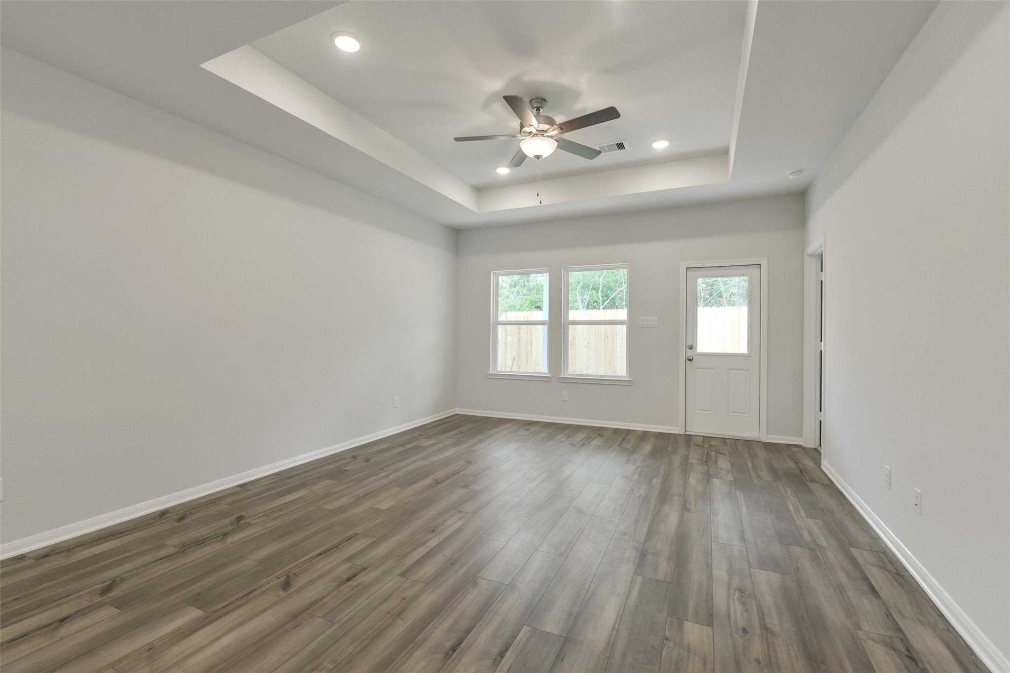 Spacious empty living room featuring tray ceiling, ceiling fan, large windows, and hardwood floors in Davidson Homes The Frio G, Conroe, Texas