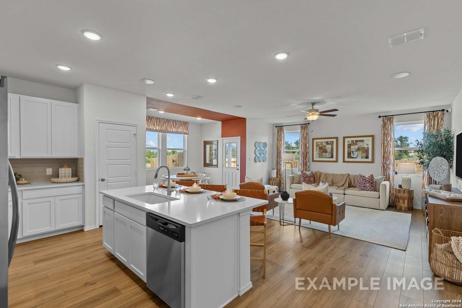 Open-concept kitchen with white cabinets, stainless dishwasher, and island bar flowing into living room with beige sofa in Davidson Homes The Daphne K, San Antonio