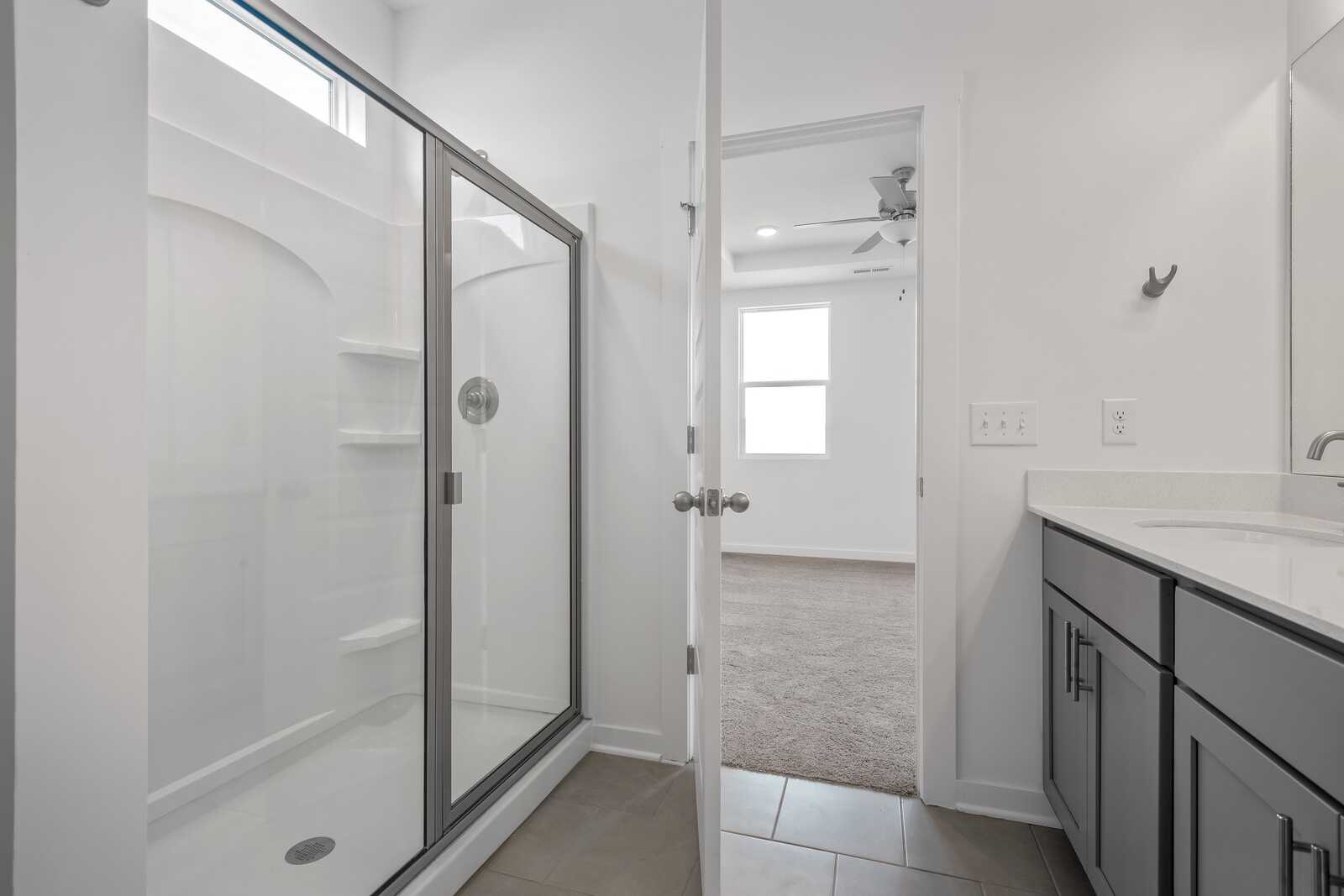 Modern master bathroom in The Asheville home design featuring frameless glass shower, dark vanity, and open bedroom view