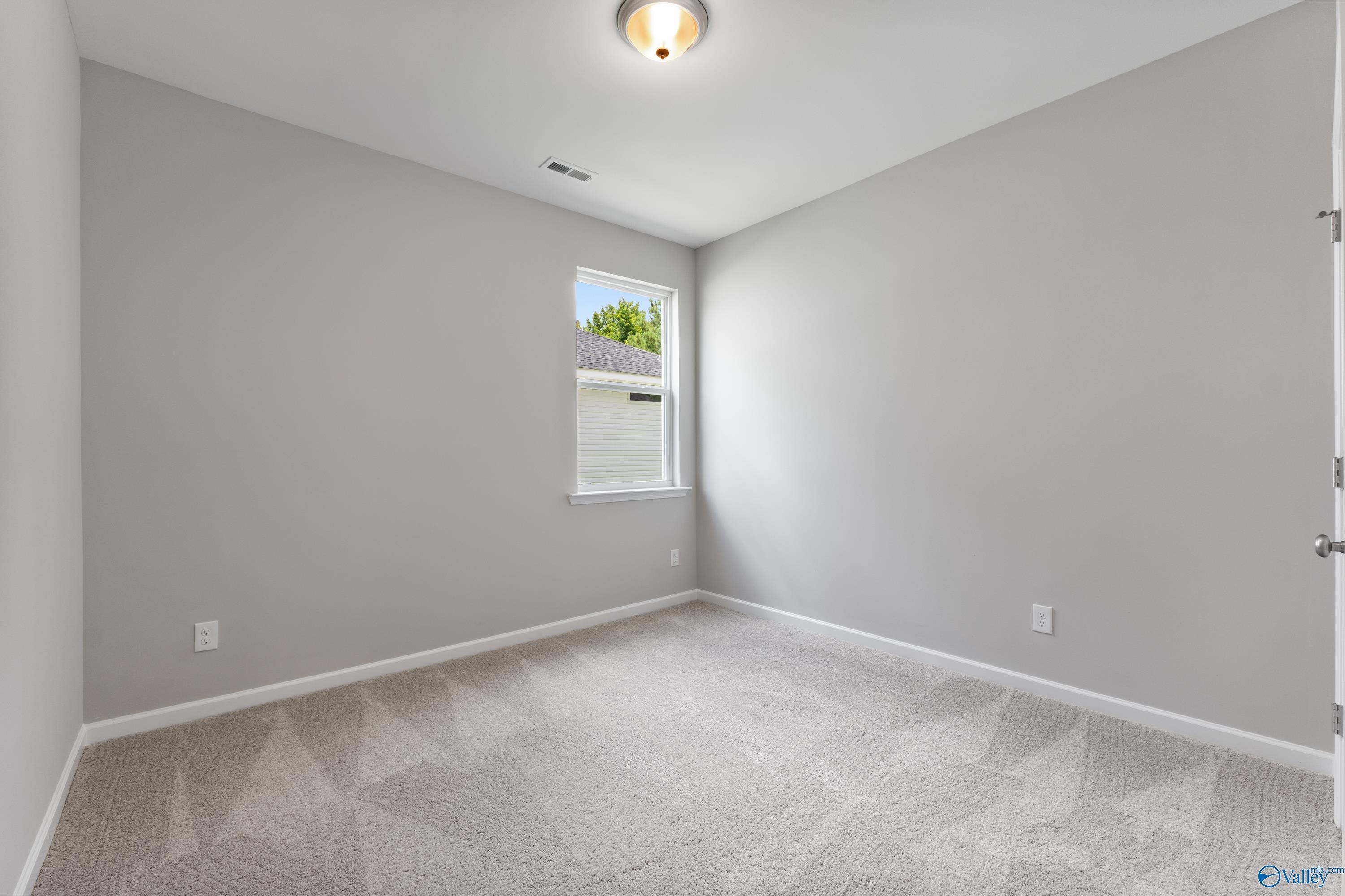 Empty bedroom with light gray walls, patterned carpet floor, and window view of trees in Davidson Homes The Phoenix, Hazel Green, Alabama