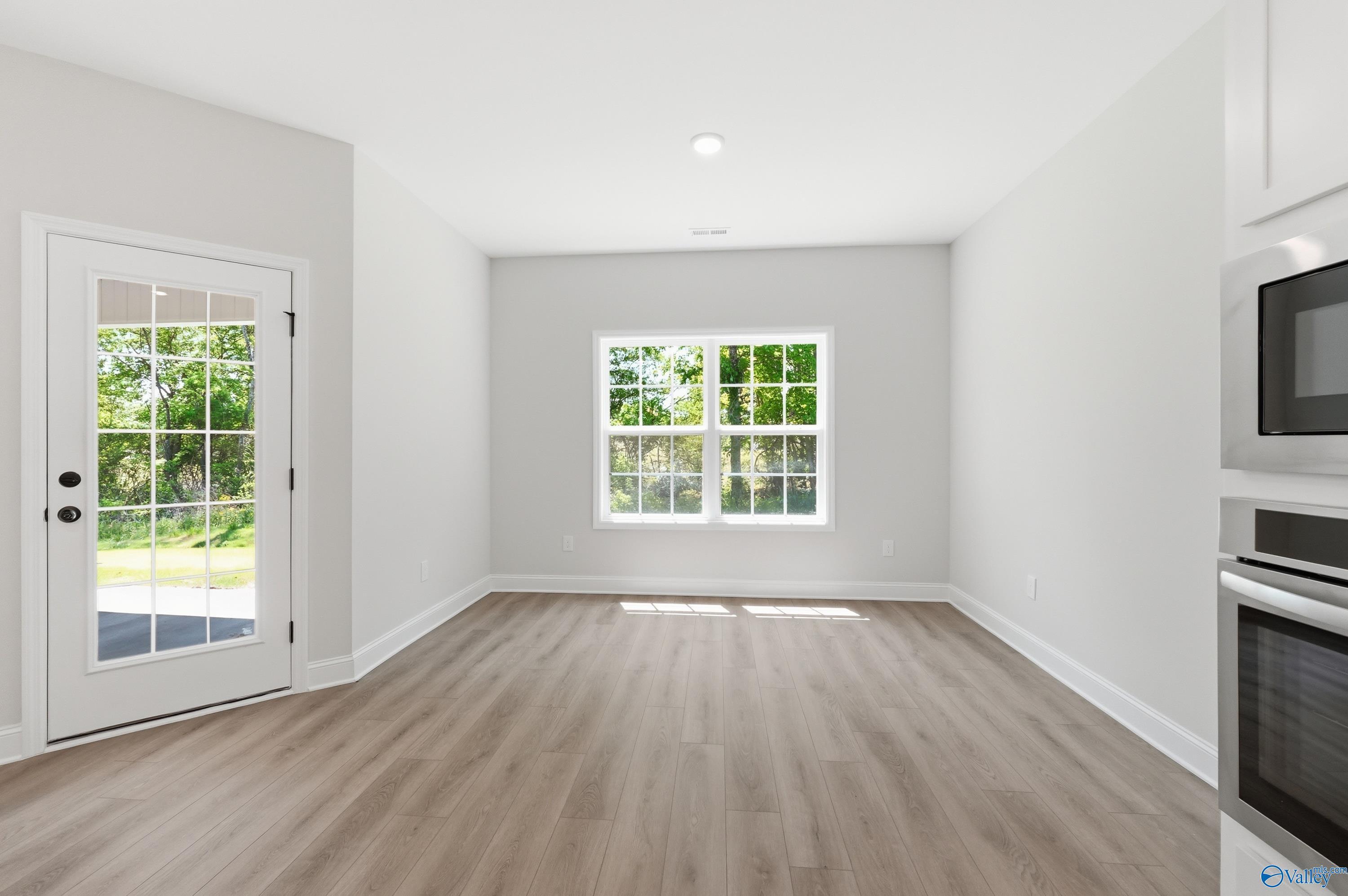 Empty living room with light hardwood floors, French door to patio, and window view of trees adjacent to kitchen in Davidson Homes The Rockford B, Athens AL