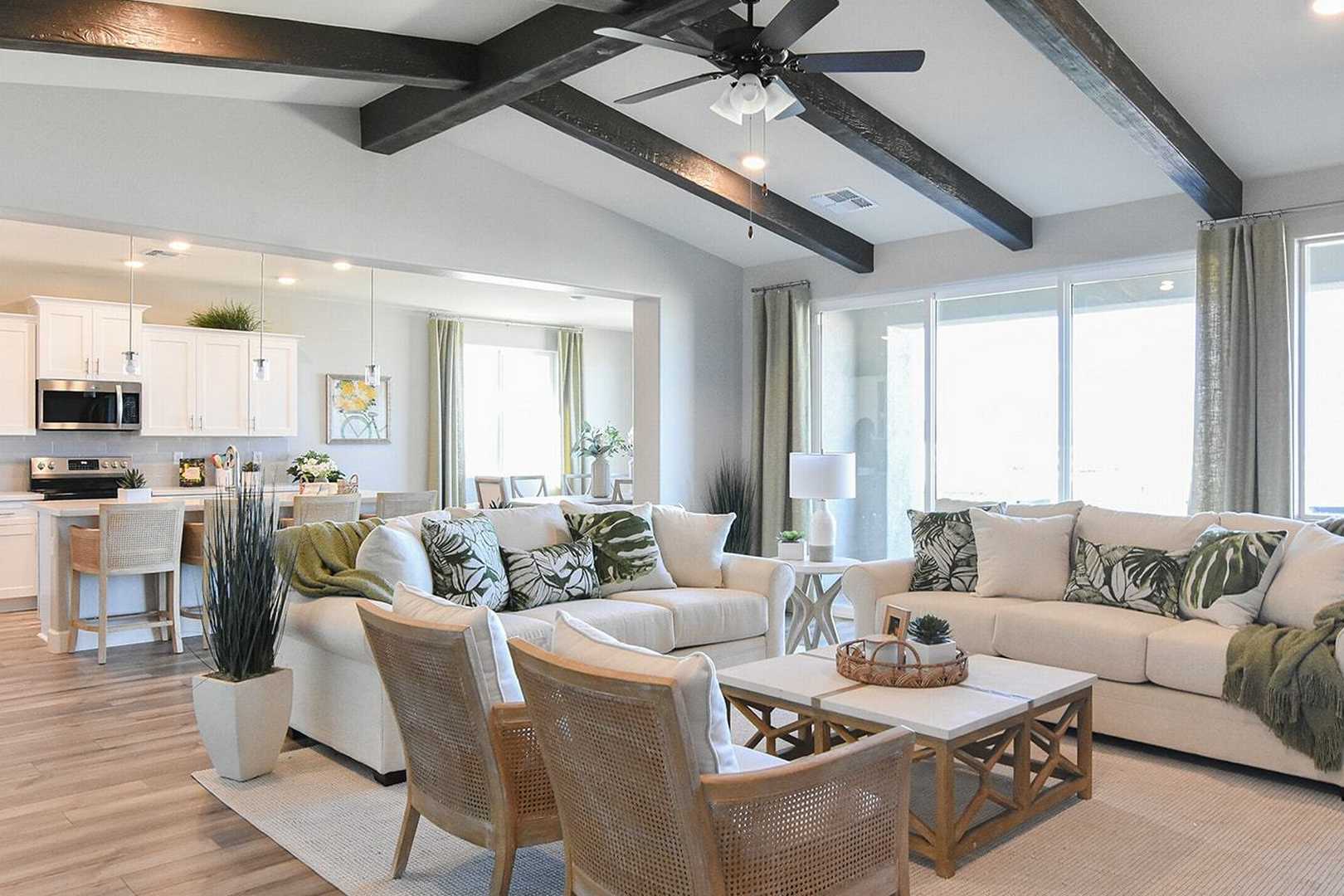 Spacious open-concept living room in The Summit B with white sectionals, wooden beams, ceiling fan, and adjacent white kitchen