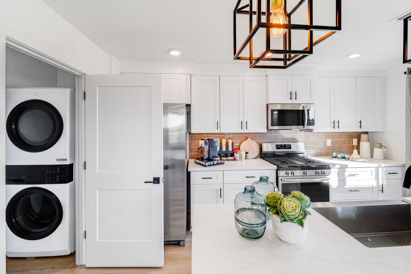Spacious laundry nook in The Wilmington home with stacked white washer dryer, adjacent modern kitchen boasting white cabinets and stainless appliances