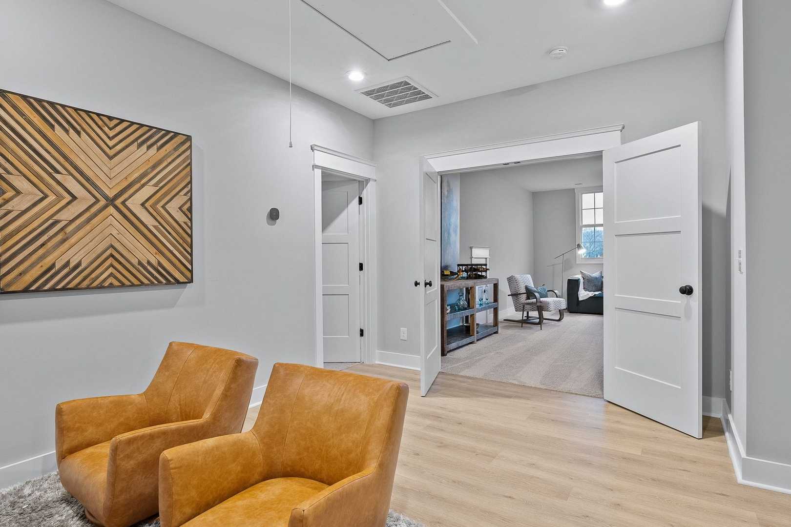 Spacious den in The Oxford home design with gray walls, orange leather armchairs, geometric wood art, and open doors to library shelves