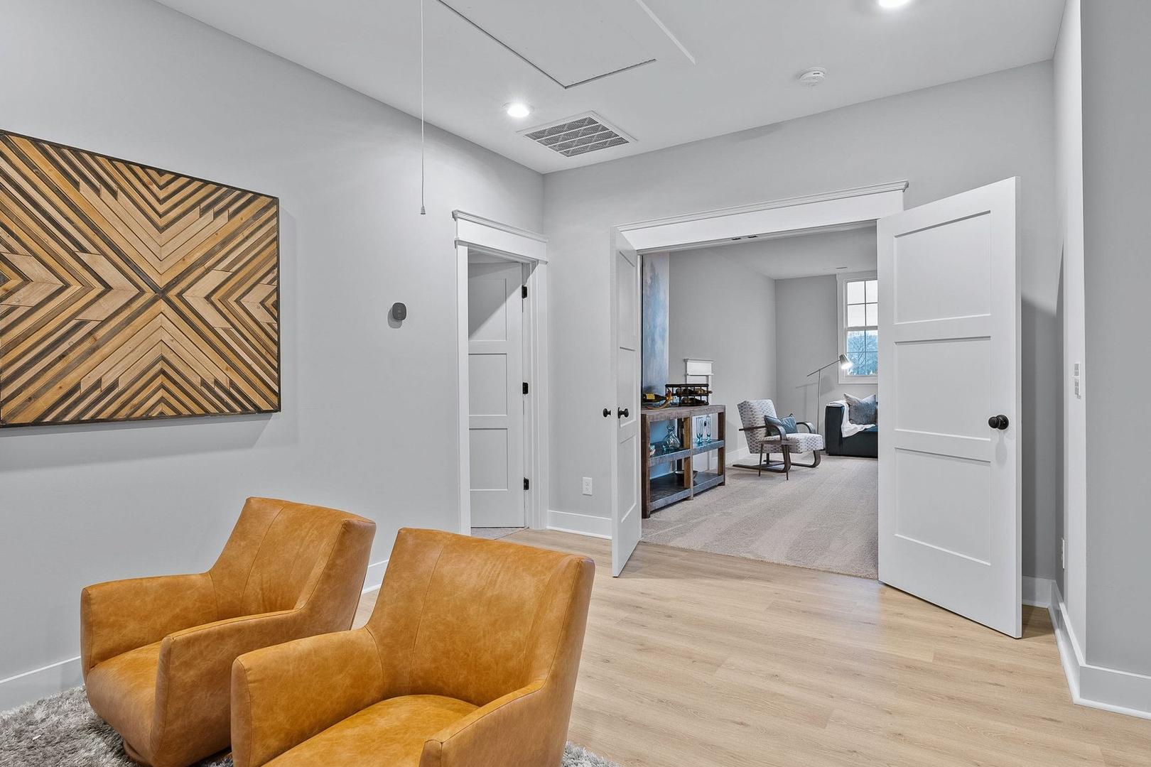 Spacious den in The Oxford home design with gray walls, orange leather armchairs, geometric wood art, and open doors to library shelves